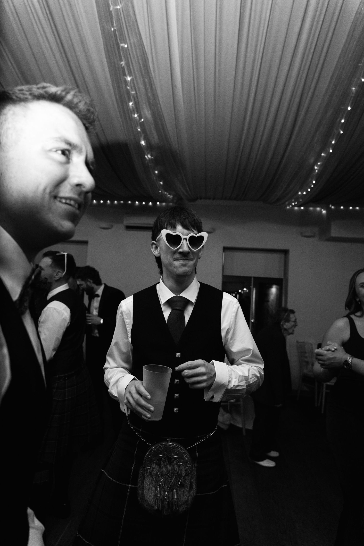 Black and white photo of wedding guests in kilts and sunglasses enjoying the reception.
