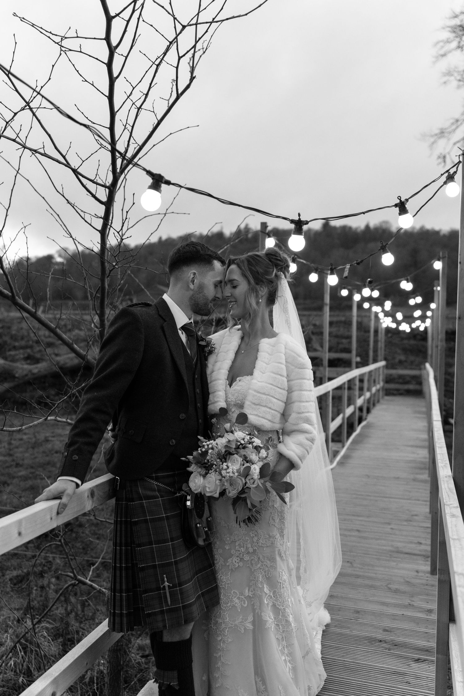 Wedding couple poses on wooden walkway with string lights and winter trees in background.