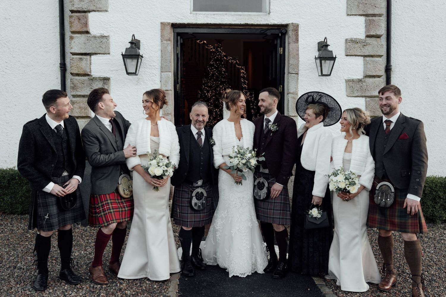 Wedding celebration outside white stone building with mix of formal wear and Scottish dress.