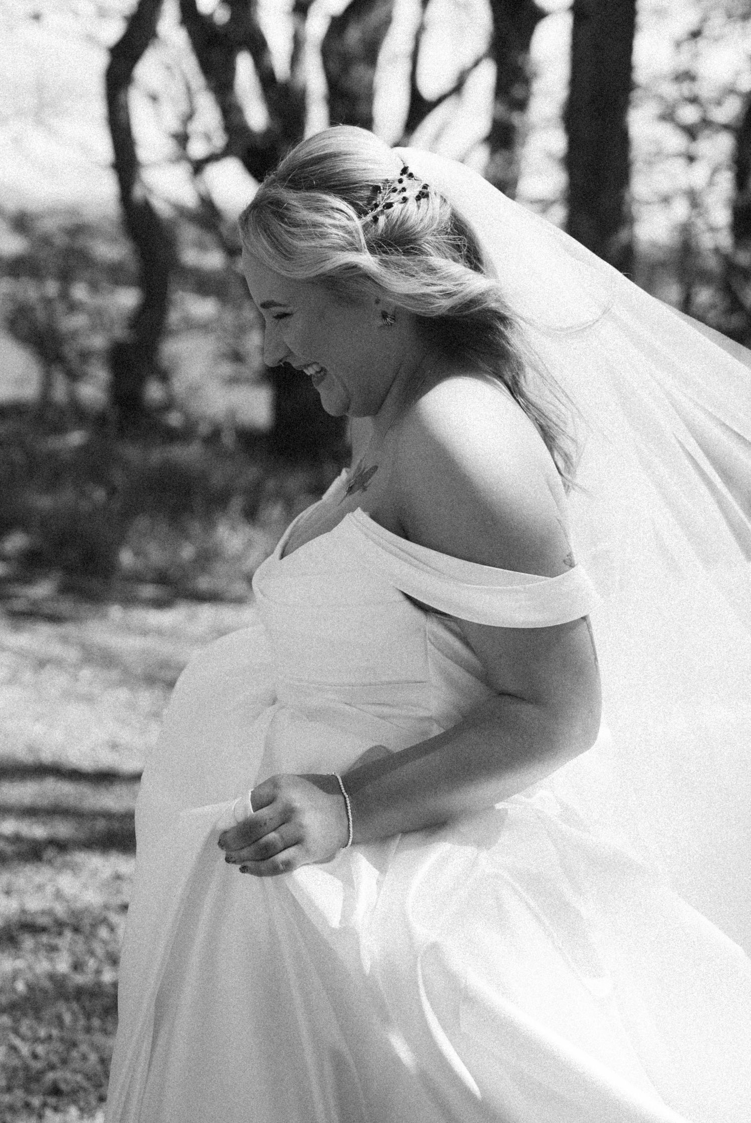 Joyful bride in off-shoulder white wedding gown and veil laughing outdoors in black and white photo.