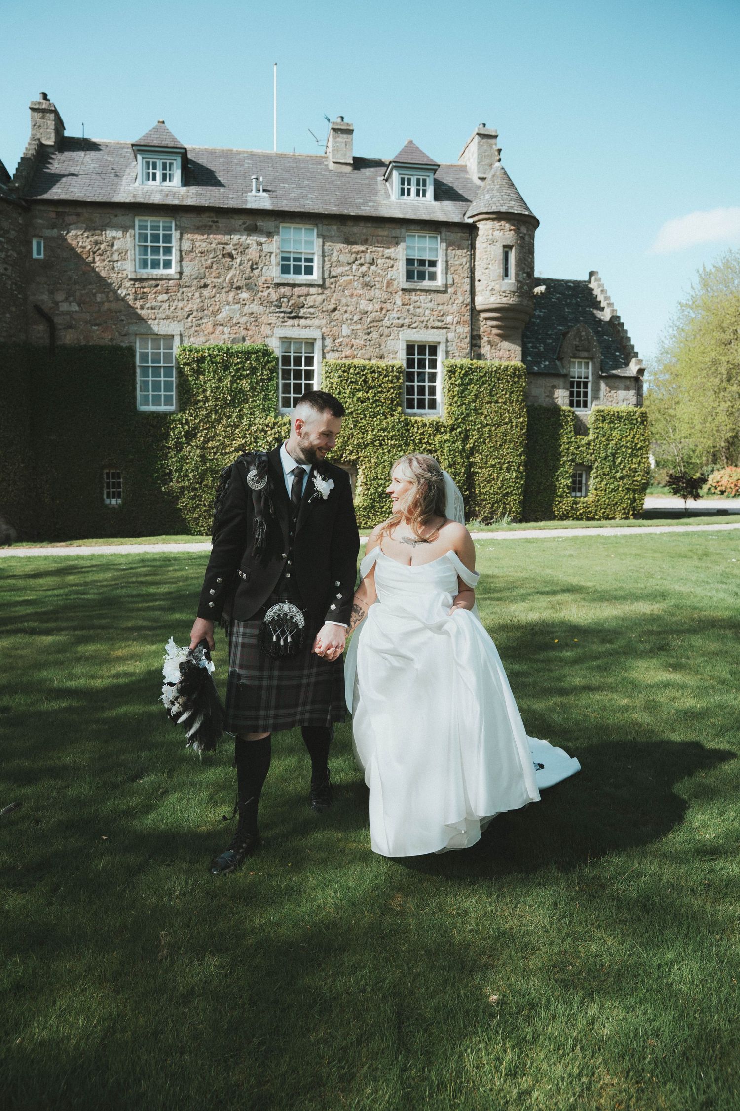 Bride and kilt-wearing groom walk hand in hand on lawn before grand ivy-covered Scottish castle.