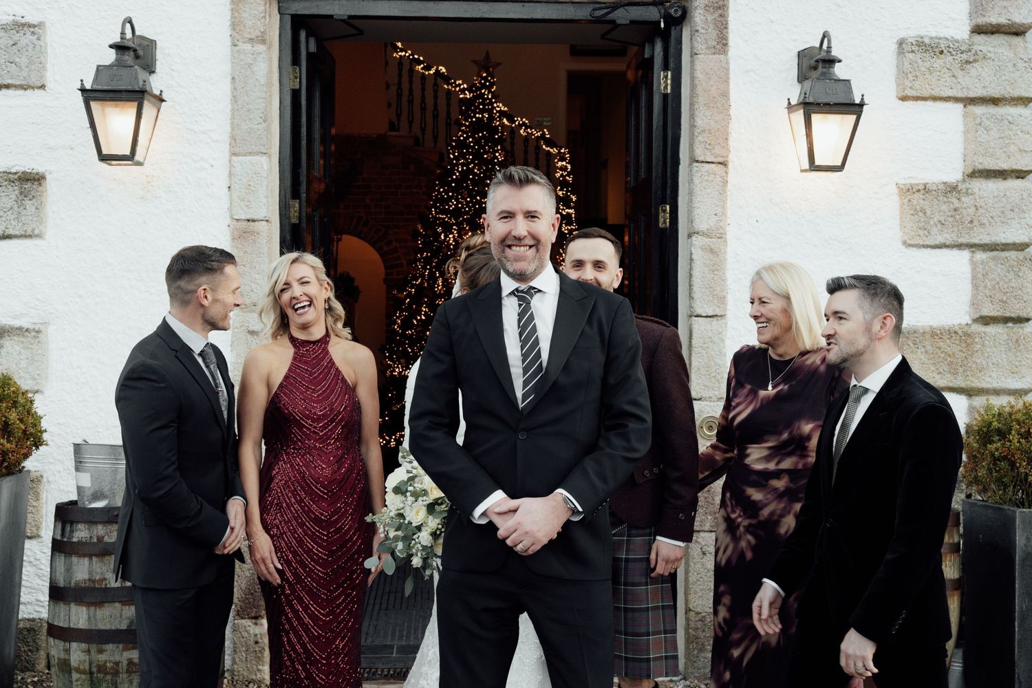 Wedding guests and bridal party gather outside a rustic venue entrance with decorative lighting.