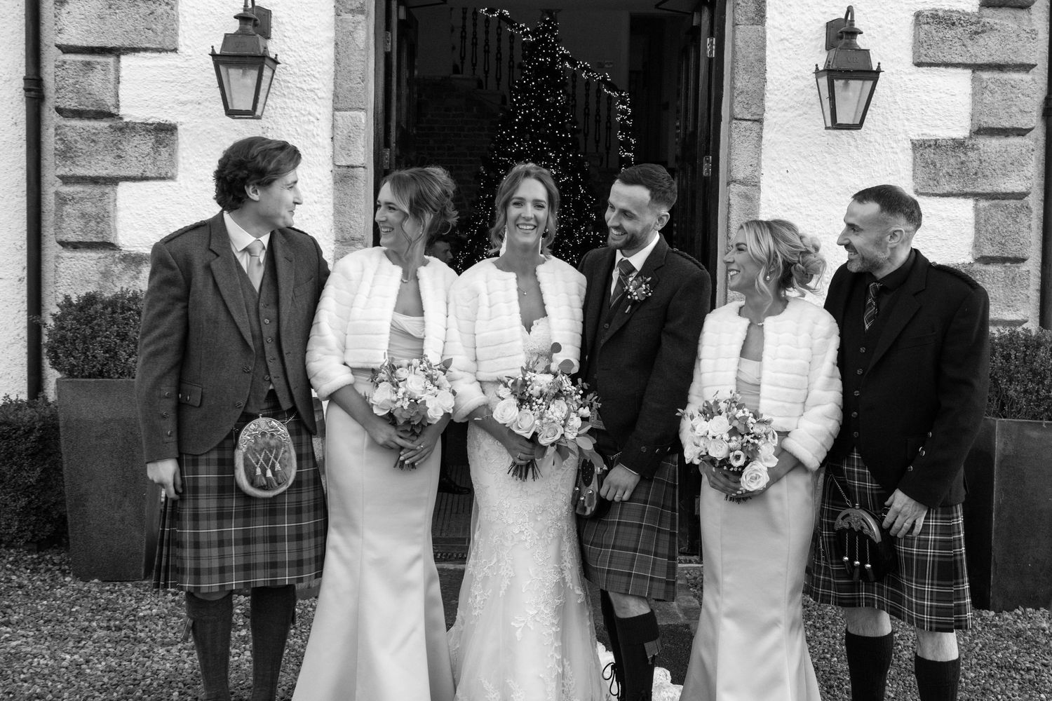 Wedding party members in kilts and white dresses pose for formal photos outside venue.