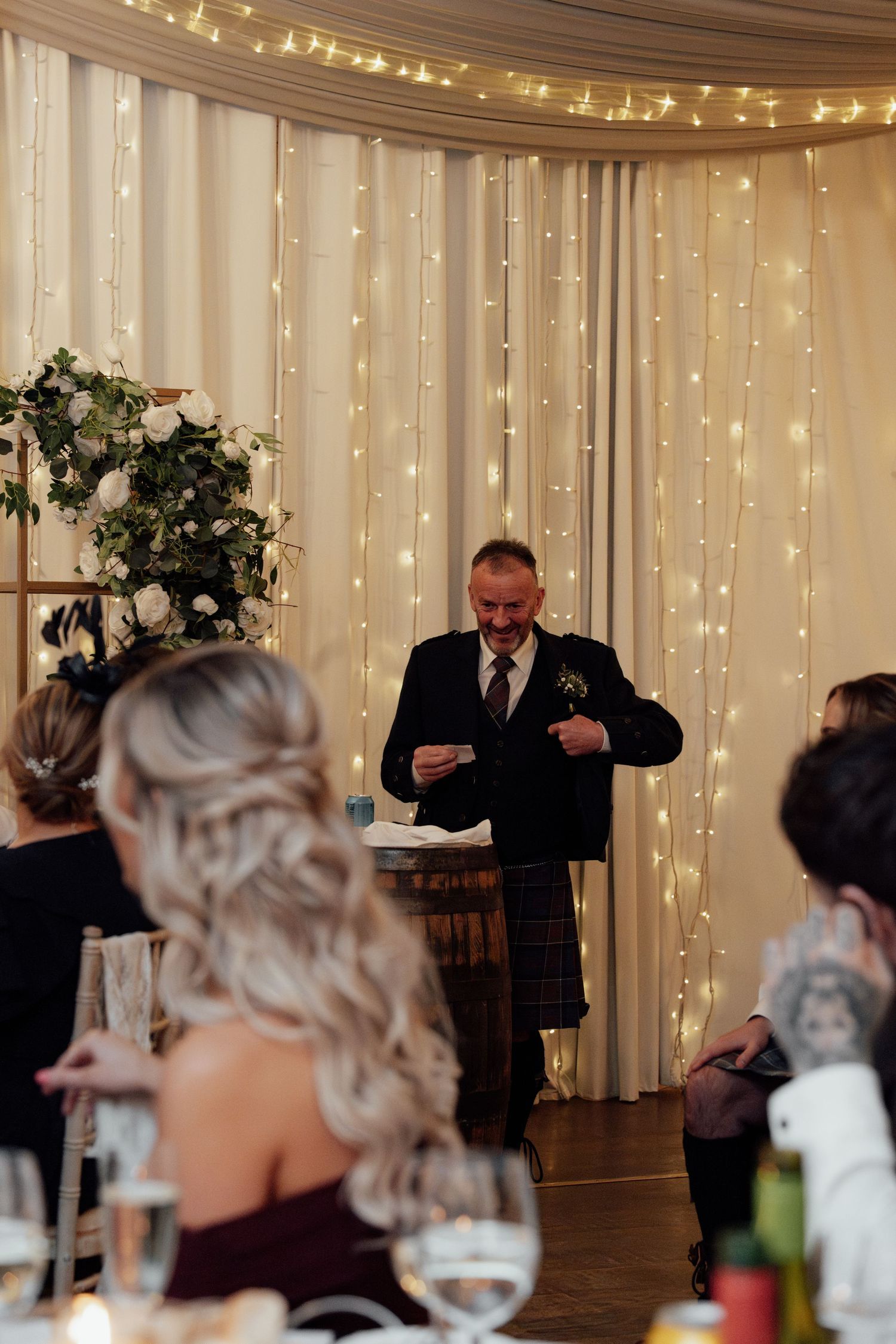 Wedding reception speech being given in an elegant room with fairy light backdrop and floral arrangements.
