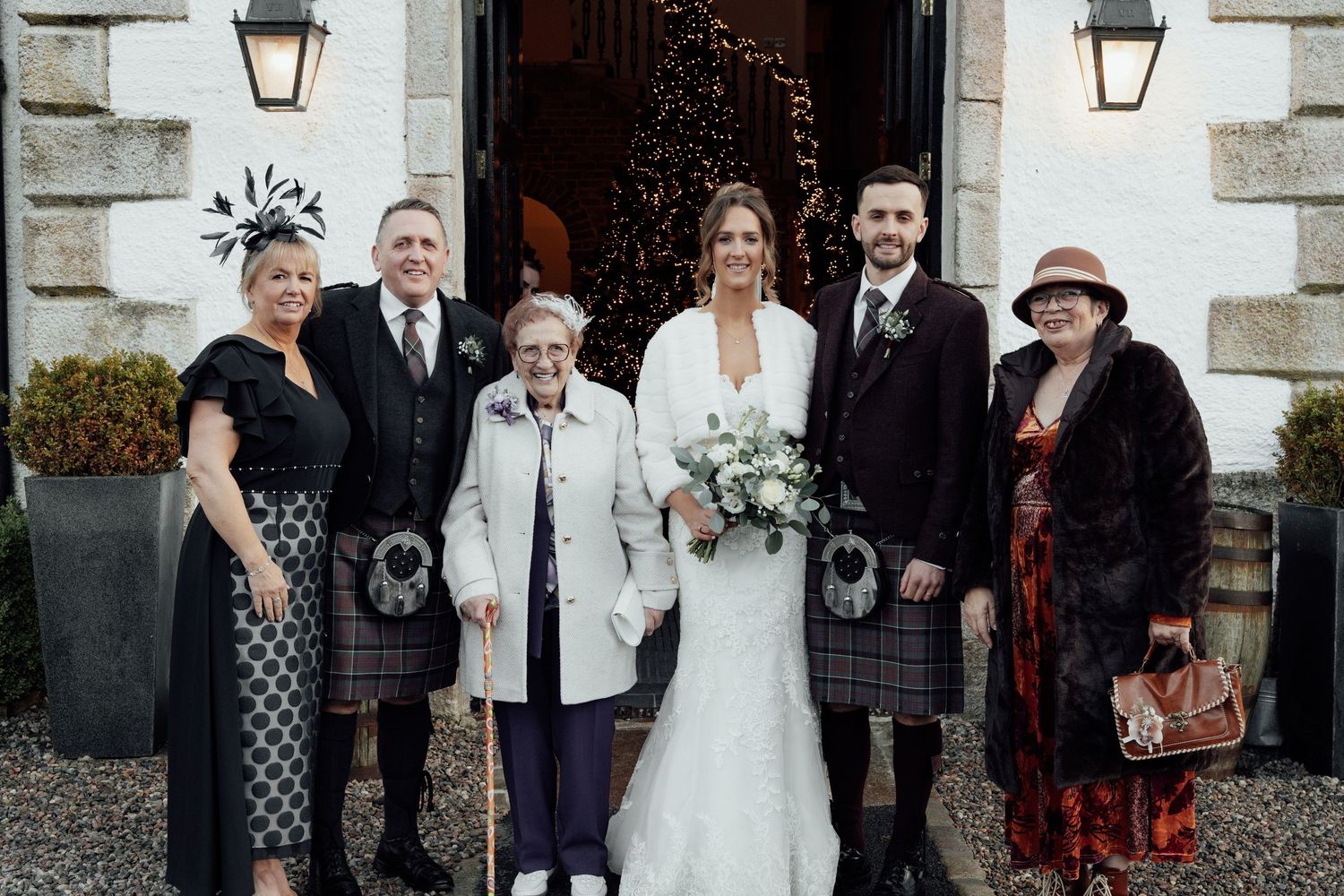 Large wedding group portrait with mix of formal wear and kilts outside stone building entrance.