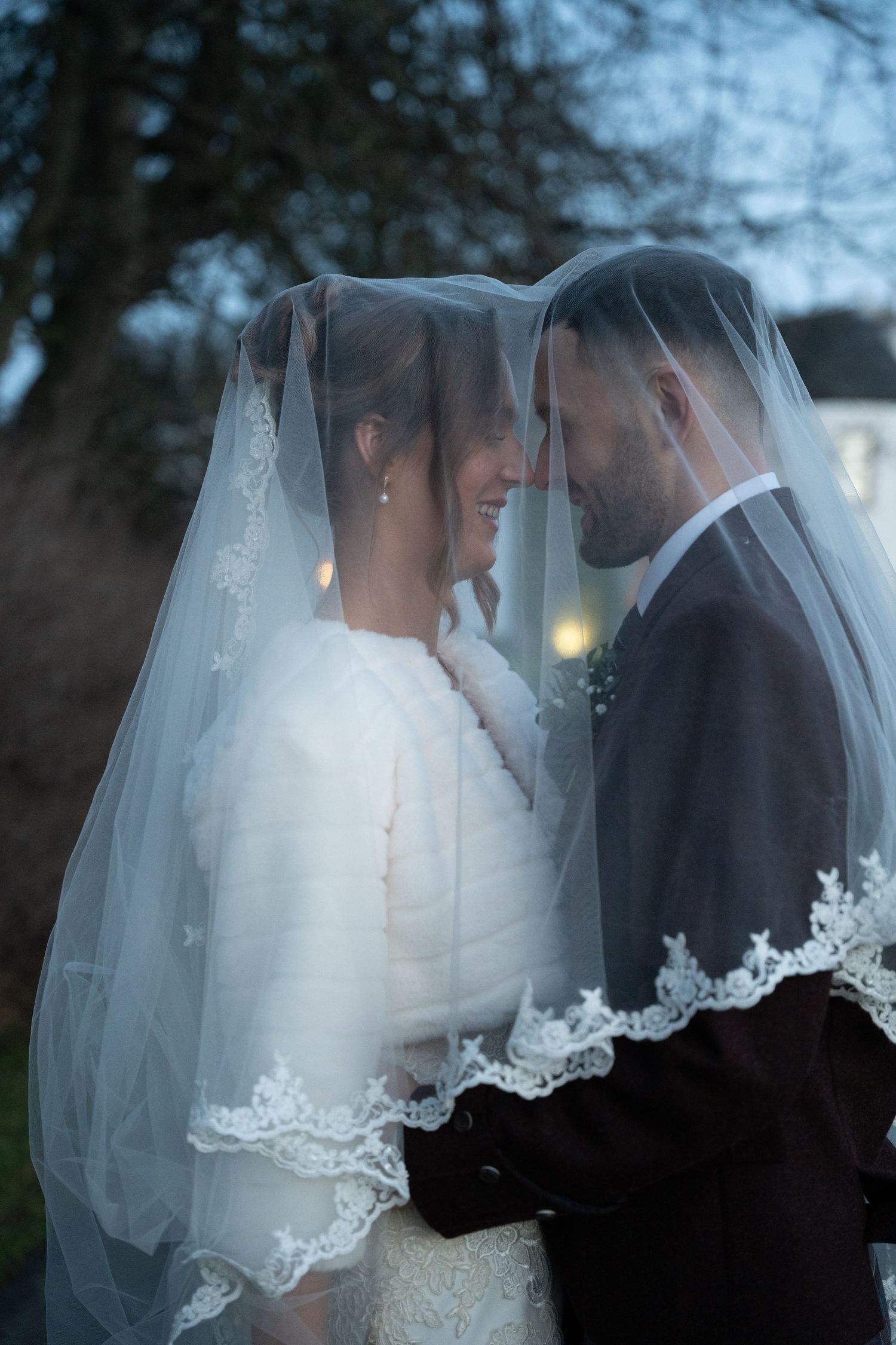 Romantic wedding portraits under a delicate lace veil in soft evening light.