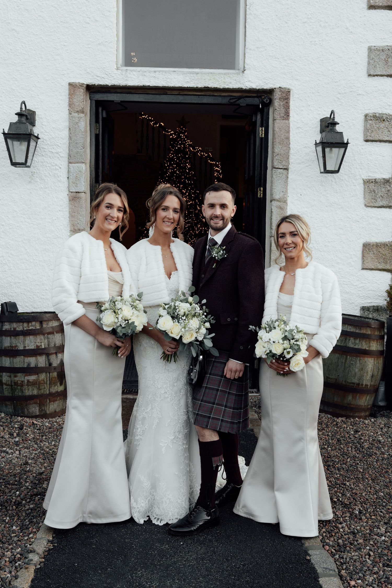Wedding group stands against white stone wall with wooden barrels and lantern decorations.