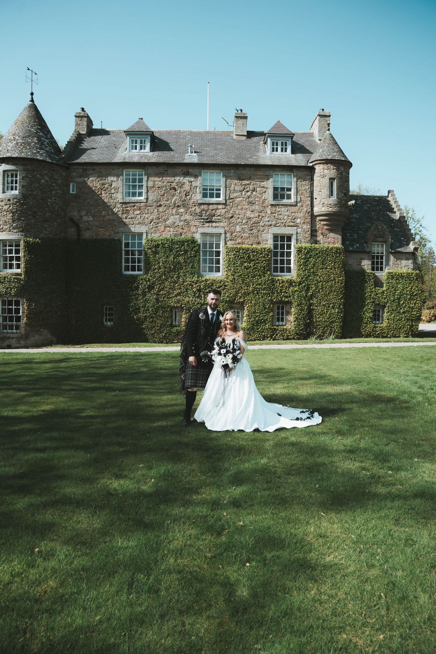 Bride and groom pose on lush green lawn before a grand ivy-covered Scottish castle on a sunny wedding day.