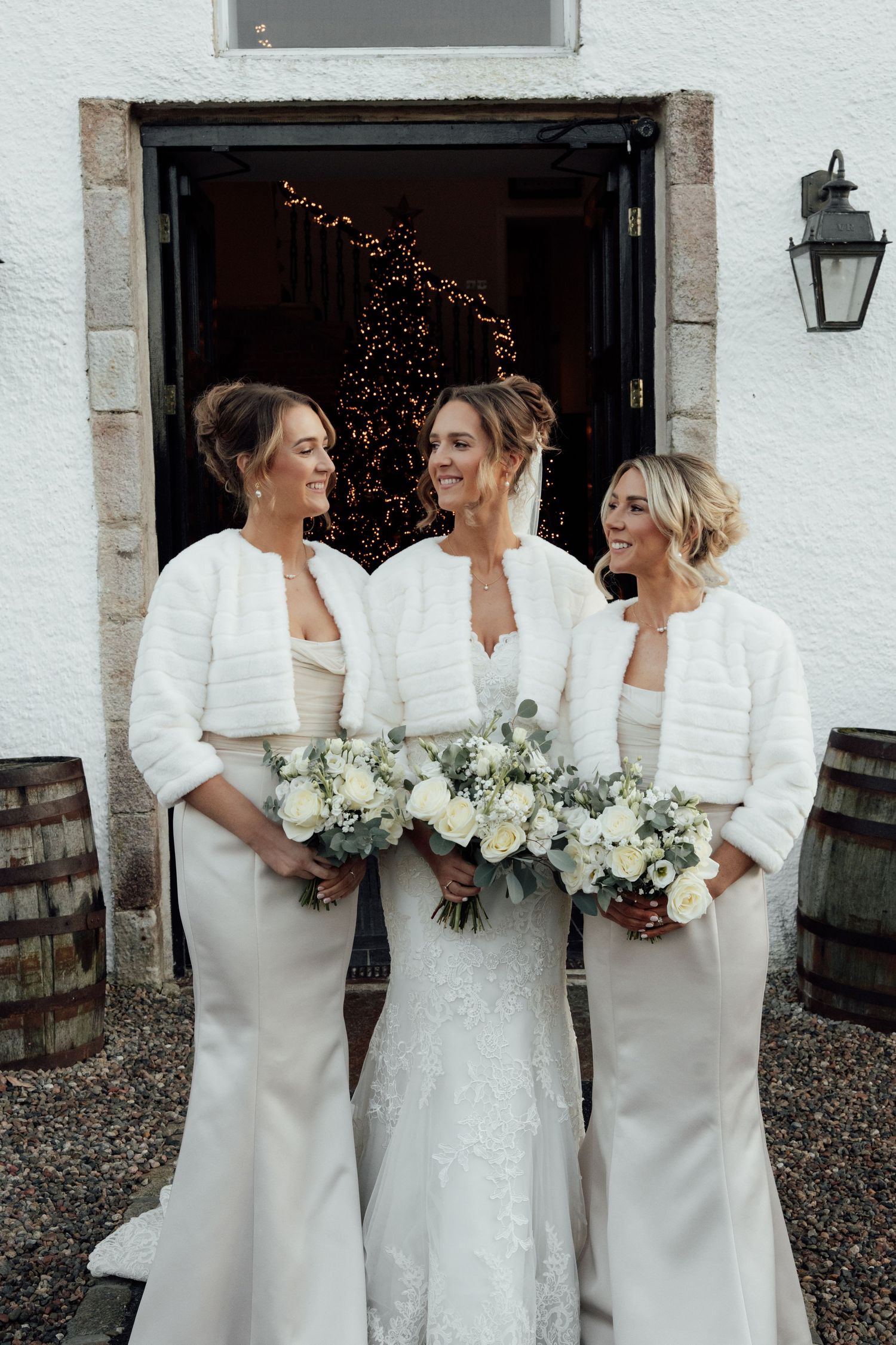 Three attendants in elegant white dresses and shawls hold white floral bouquets while sharing a joyful moment.