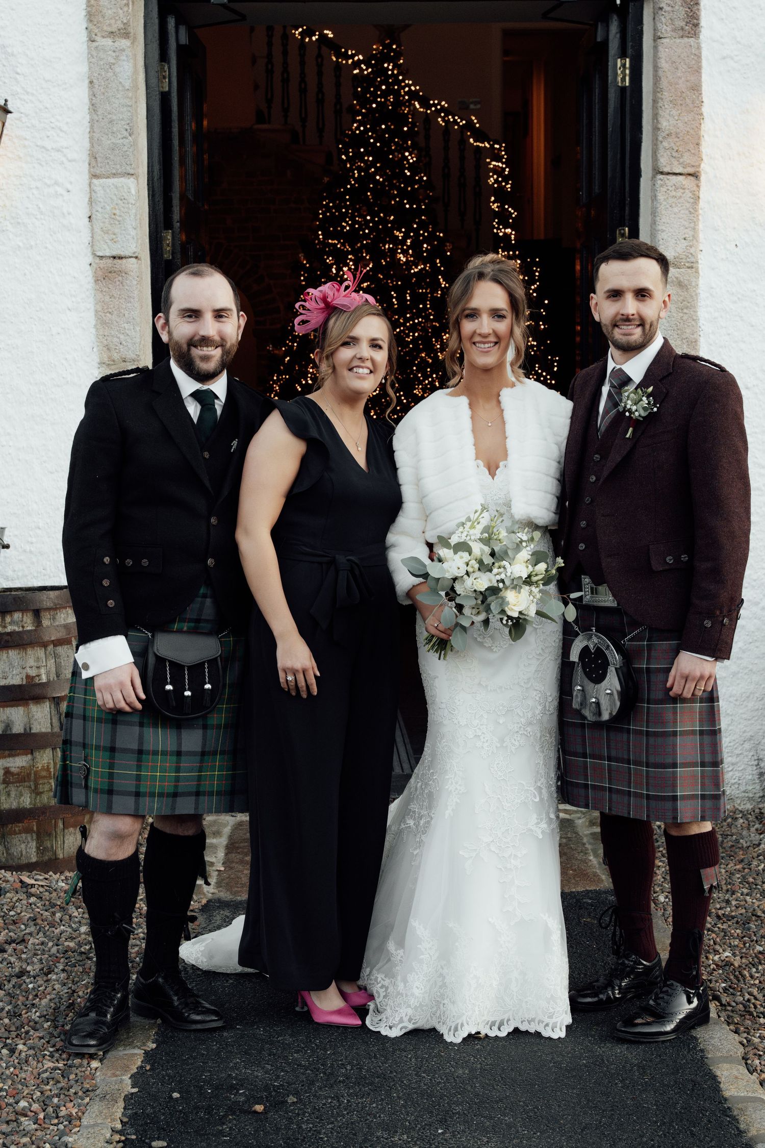 Wedding party members pose in mix of kilts, formal dresses, and bridal gown outside venue entrance.