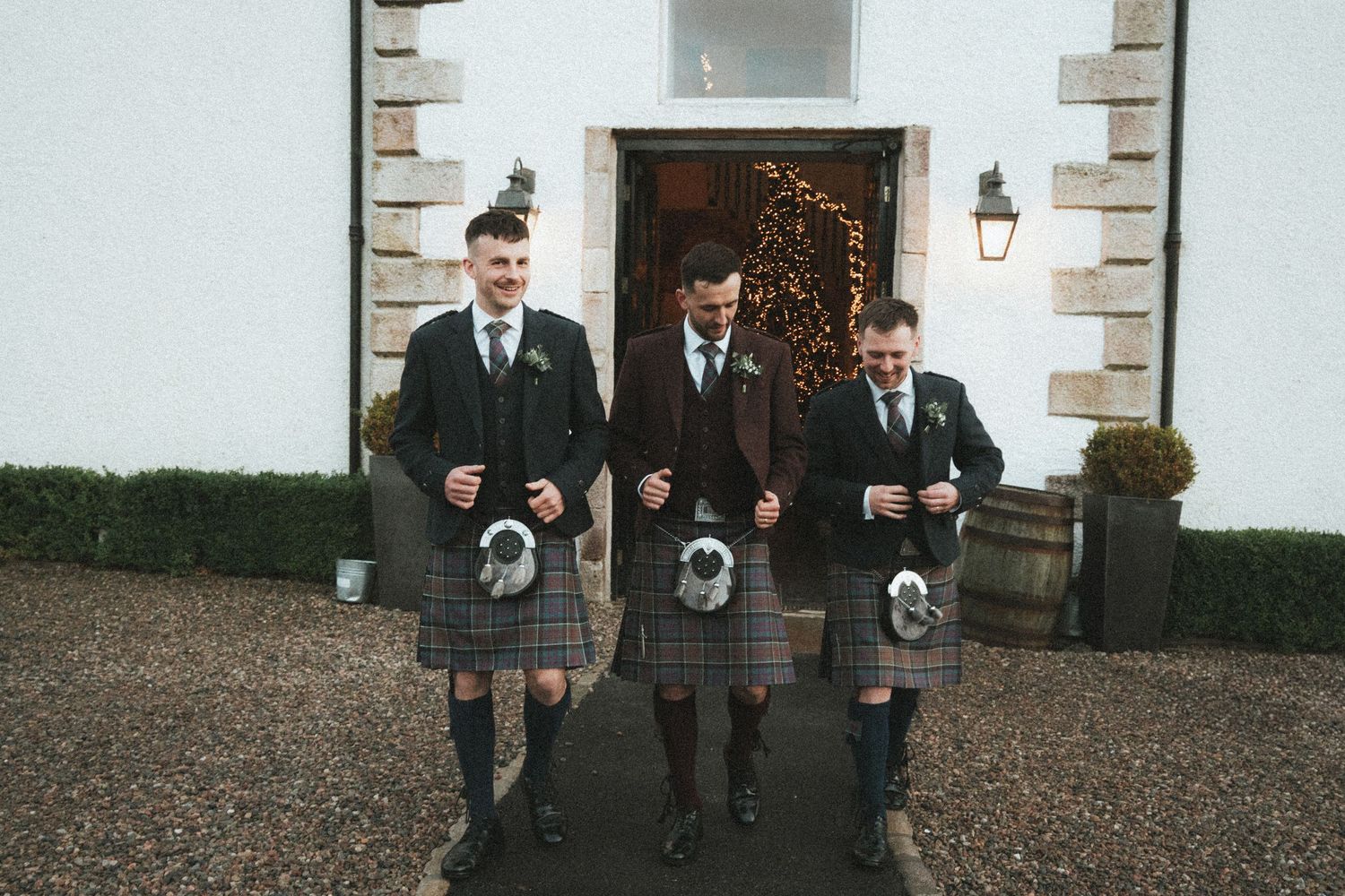 Three groomsmen in traditional Scottish dress descend stone steps at venue entrance.