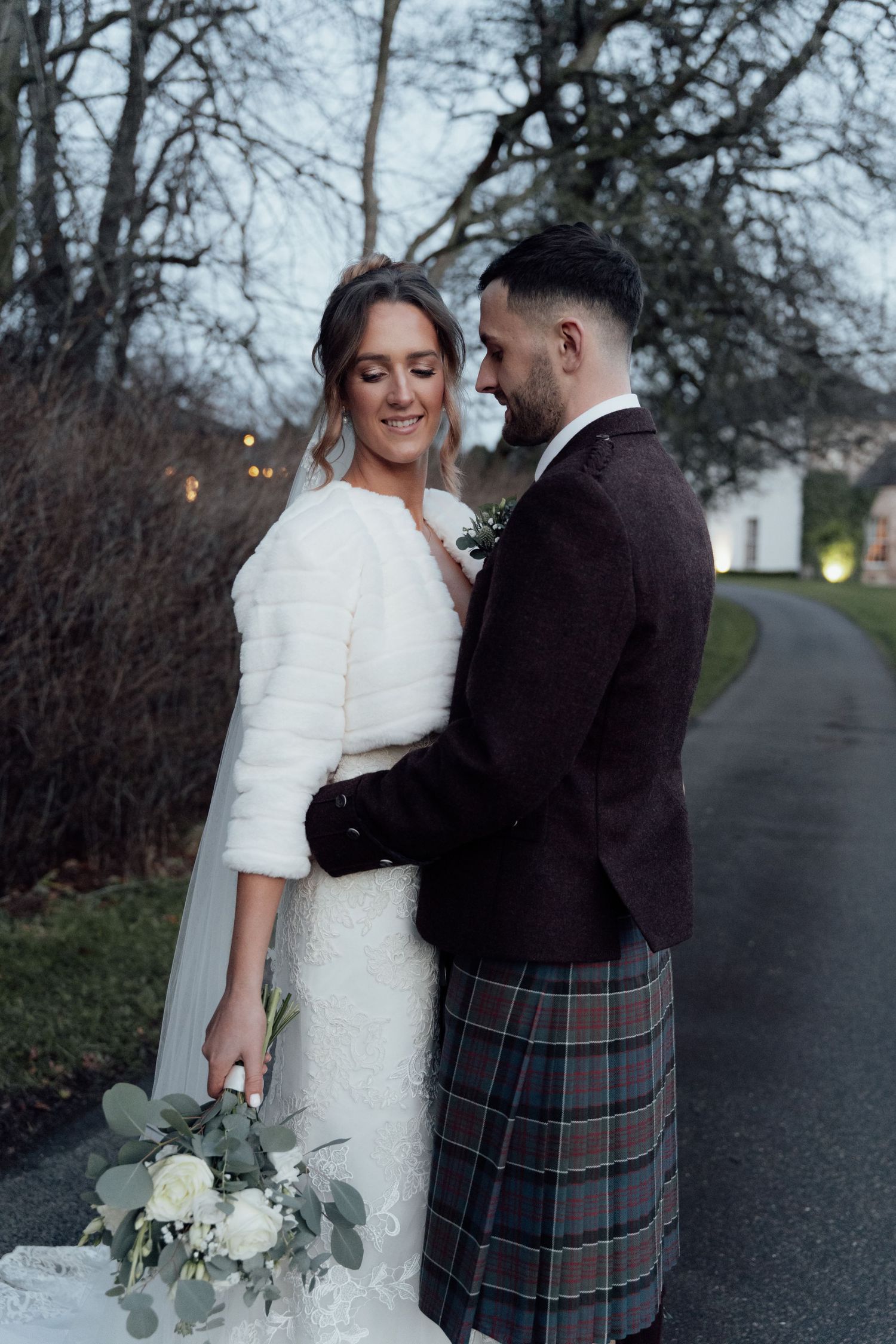 Newlyweds pose together on a path with the bride holding a bouquet and groom wearing a traditional Scottish kilt.