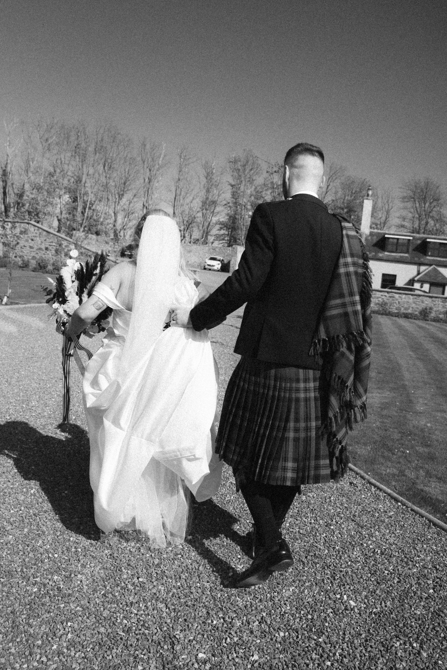 Bride and groom walking hand in hand away from camera, groom wearing traditional Scottish kilt.
