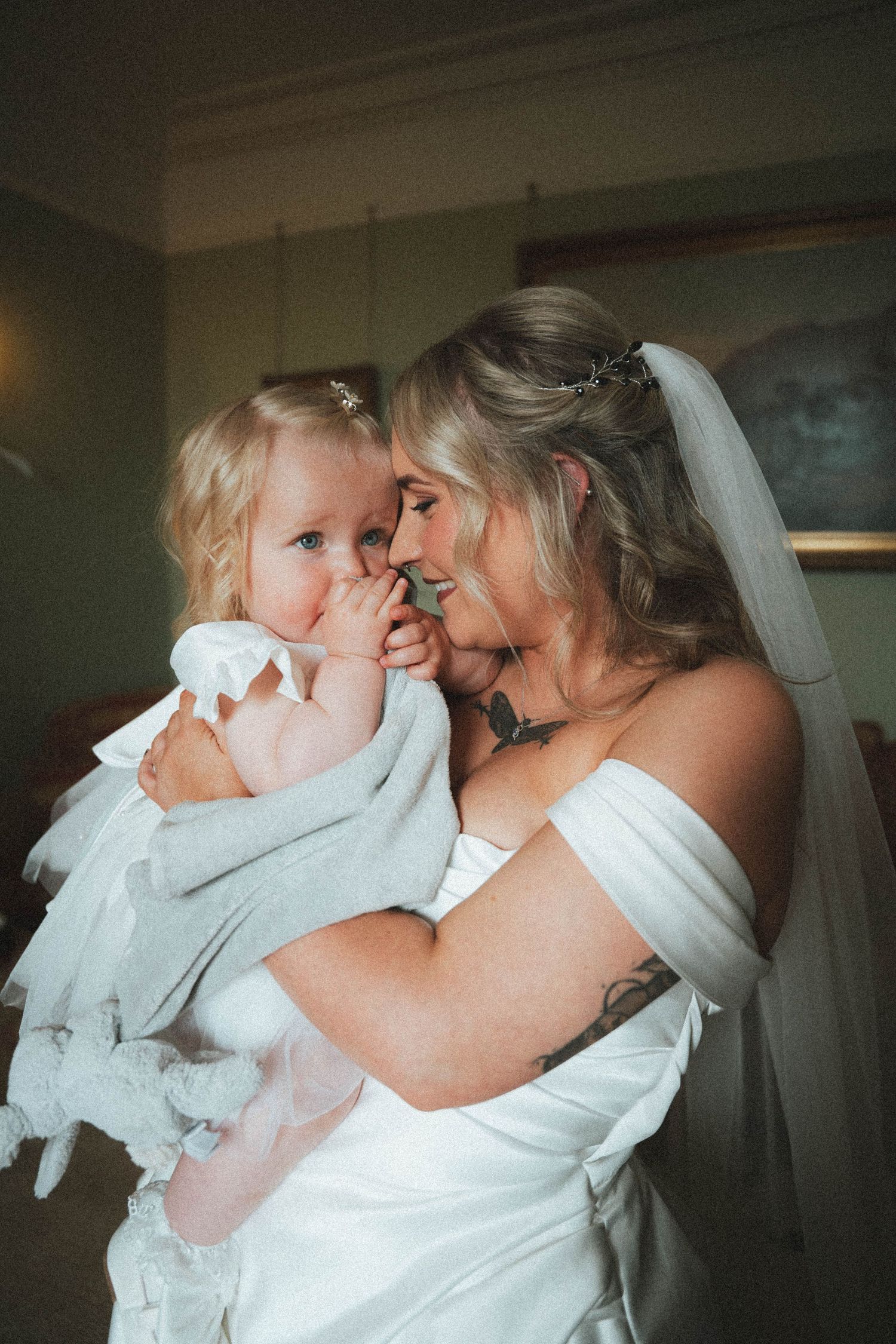 Bride in white off-shoulder wedding dress tenderly holding a young blonde toddler girl in a softly lit room.