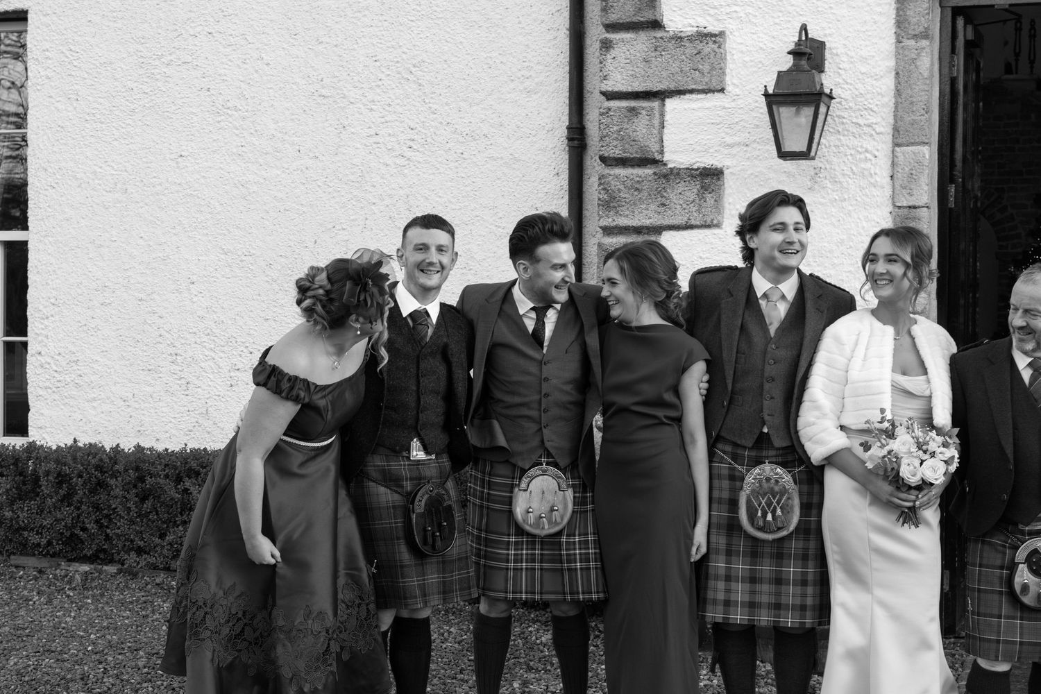 Wedding party members in Scottish formal wear standing near stone building entrance with lanterns.