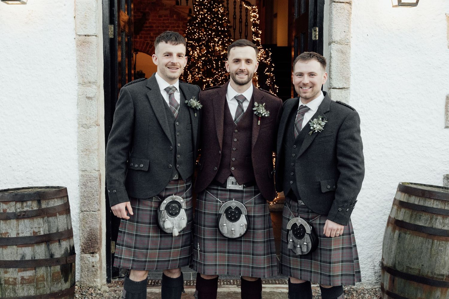 Three men in matching kilts and formal jackets stand between wooden barrels at venue entrance.