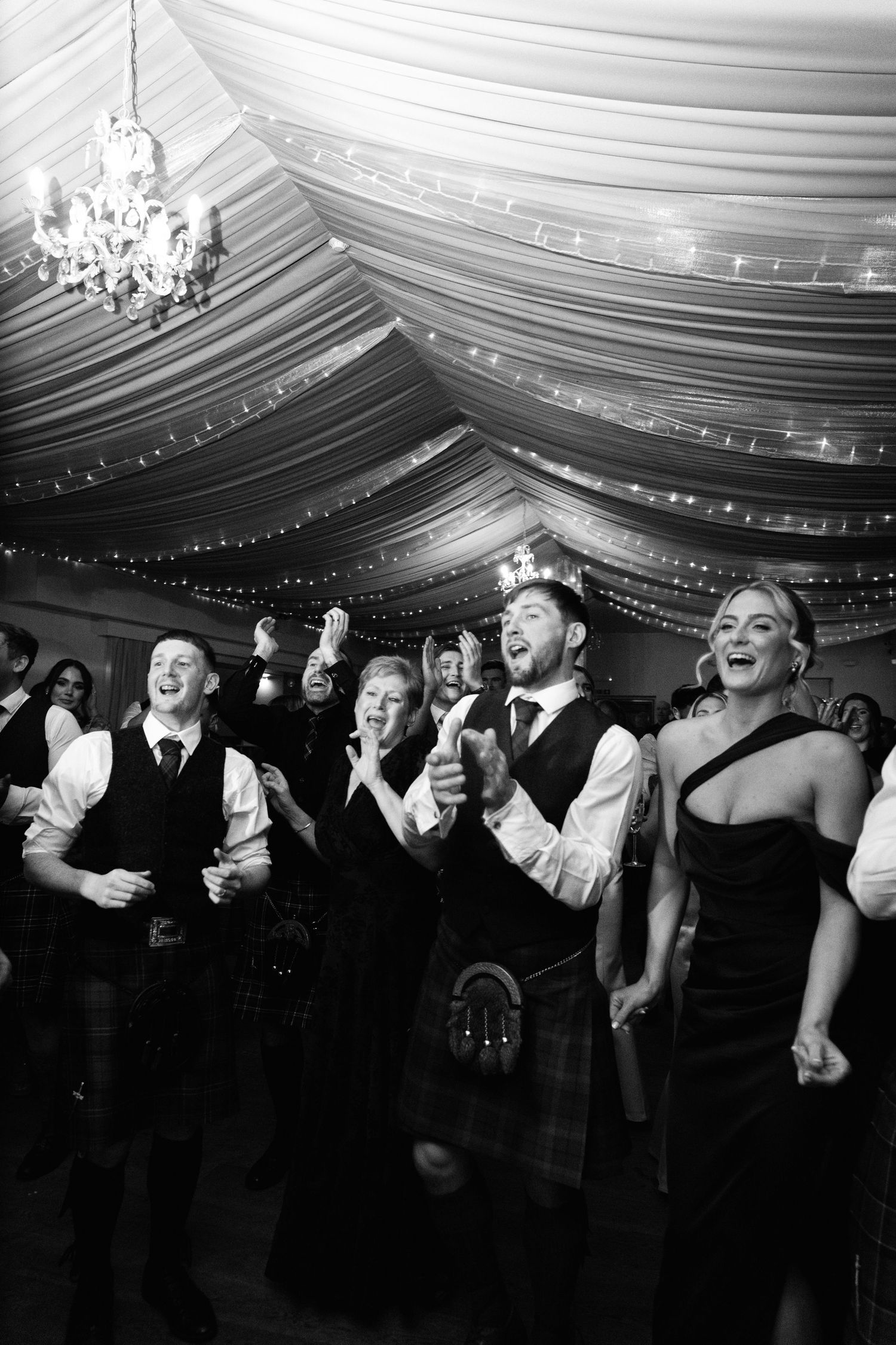 Black and white photo of wedding guests dancing and celebrating under draped tent ceiling.