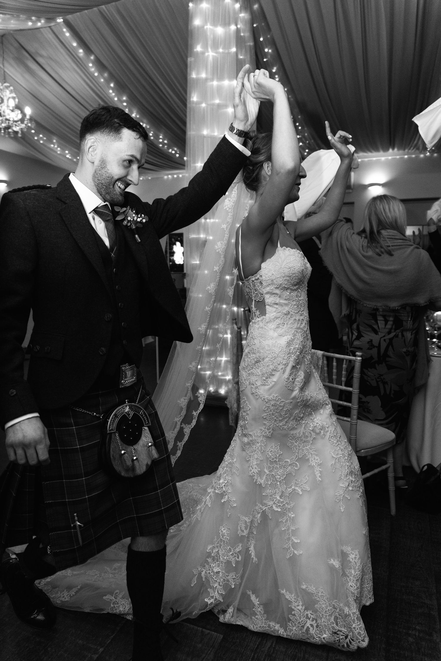 Wedding guests dance and celebrate on the dance floor under twinkling lights in a draped reception tent.