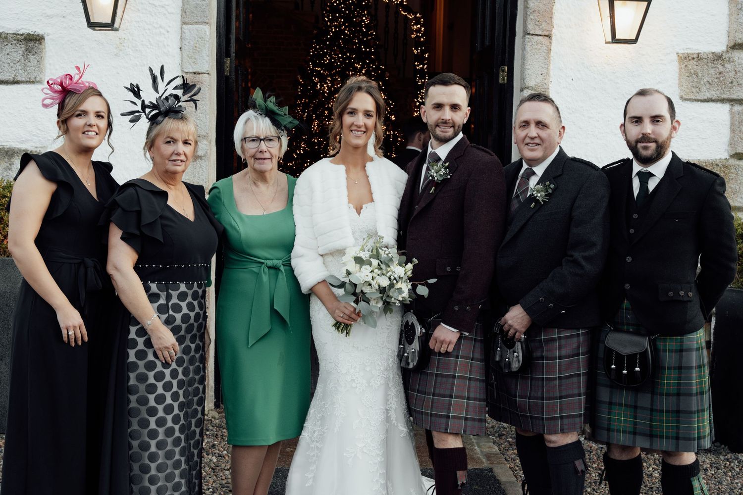 Wedding group photo featuring Scottish kilts and formal dresses outside stone building.