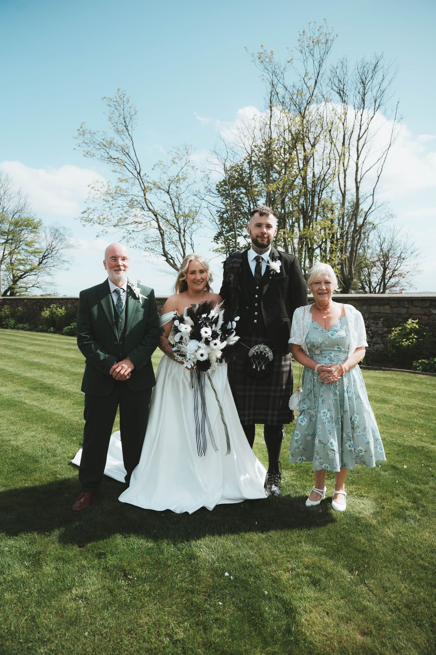 Wedding party of four posing outdoors on green lawn, groom in Scottish tartan kilt with family.