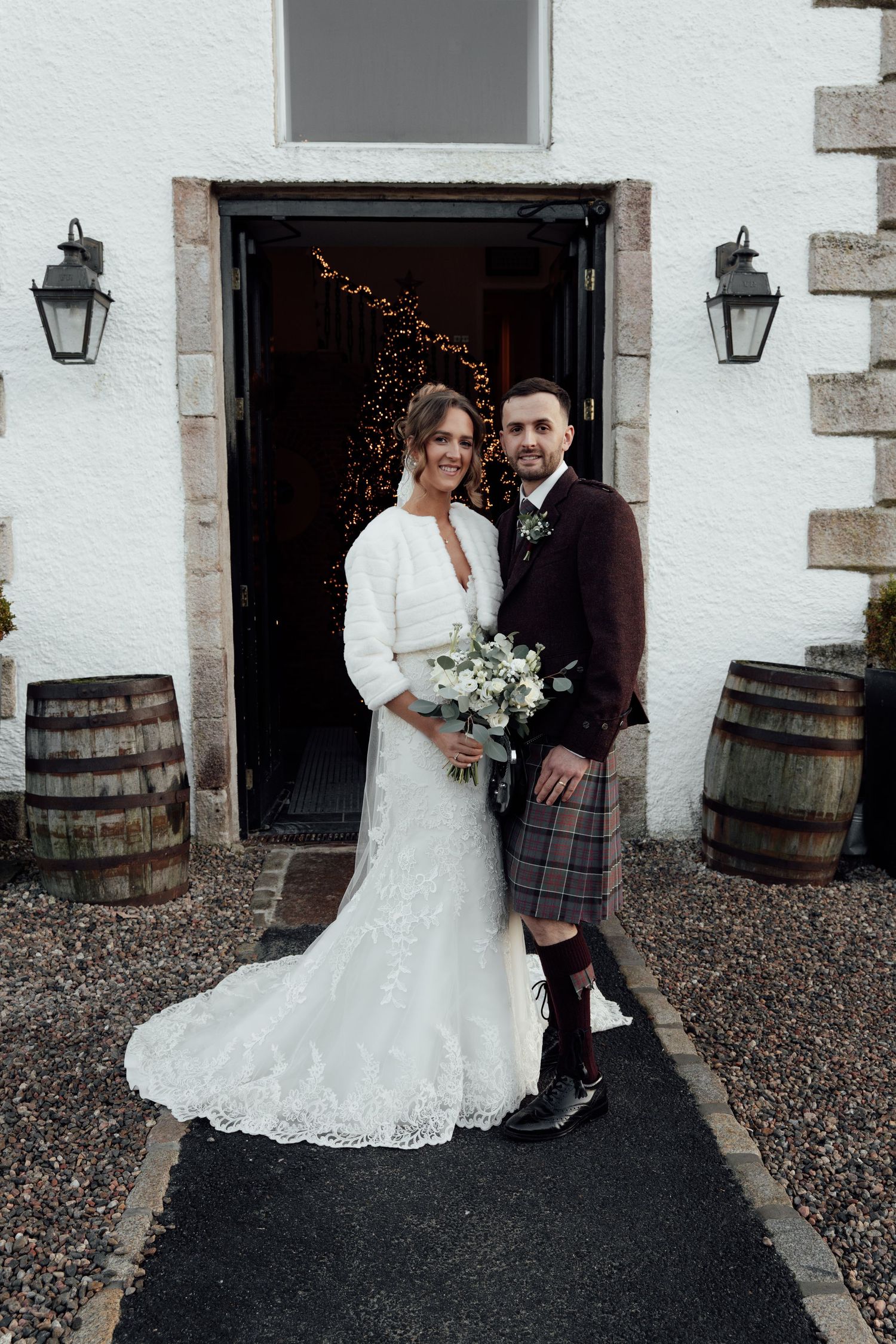 Scottish wedding couple posing by wooden barrels outside white stone building.