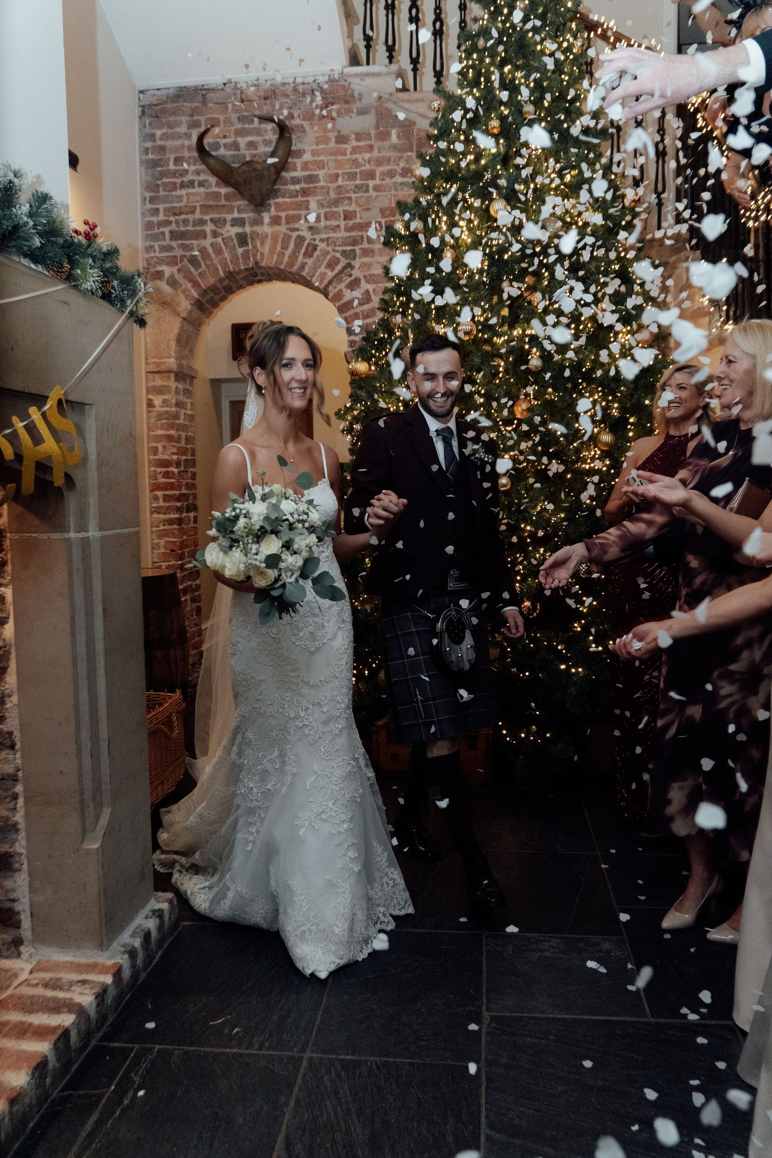 Bride in lace gown walks through shower of confetti near illuminated Christmas tree.