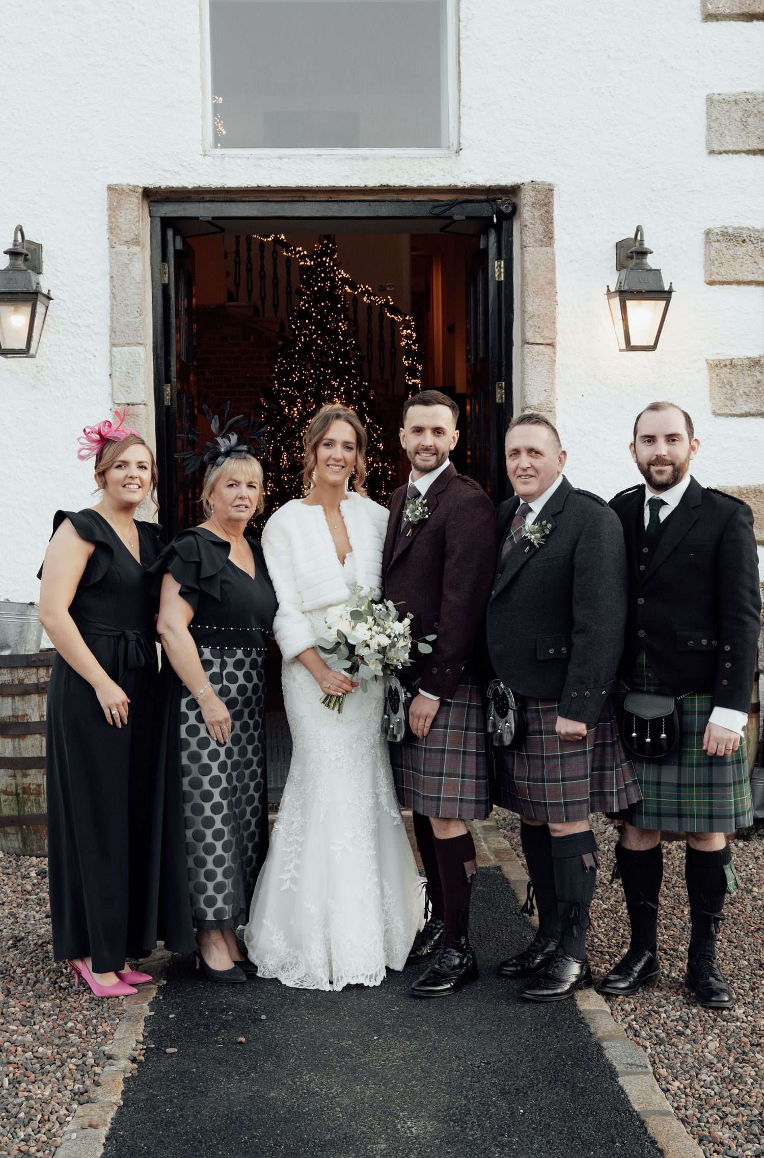 Wedding party in mix of kilts and formal dresses poses against white building exterior.