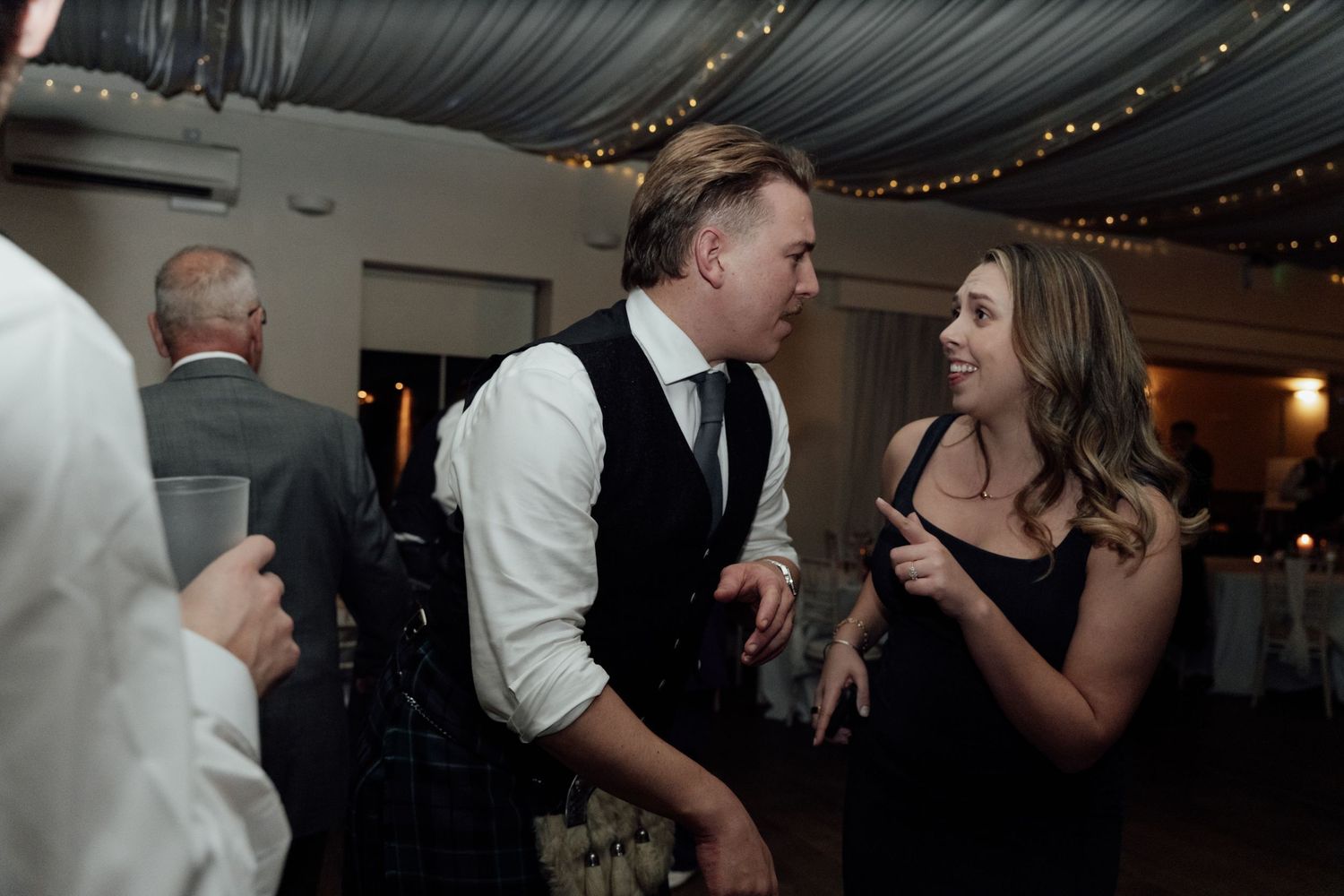 Wedding guests dancing and socializing at a reception under draped fabric ceiling with string lights.