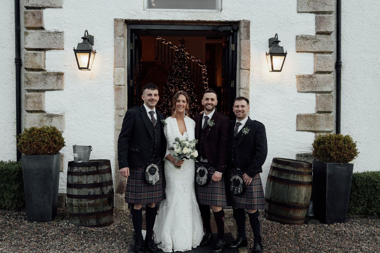 Elegant wedding party gathered outside white building with stone trim and decorative barrels.