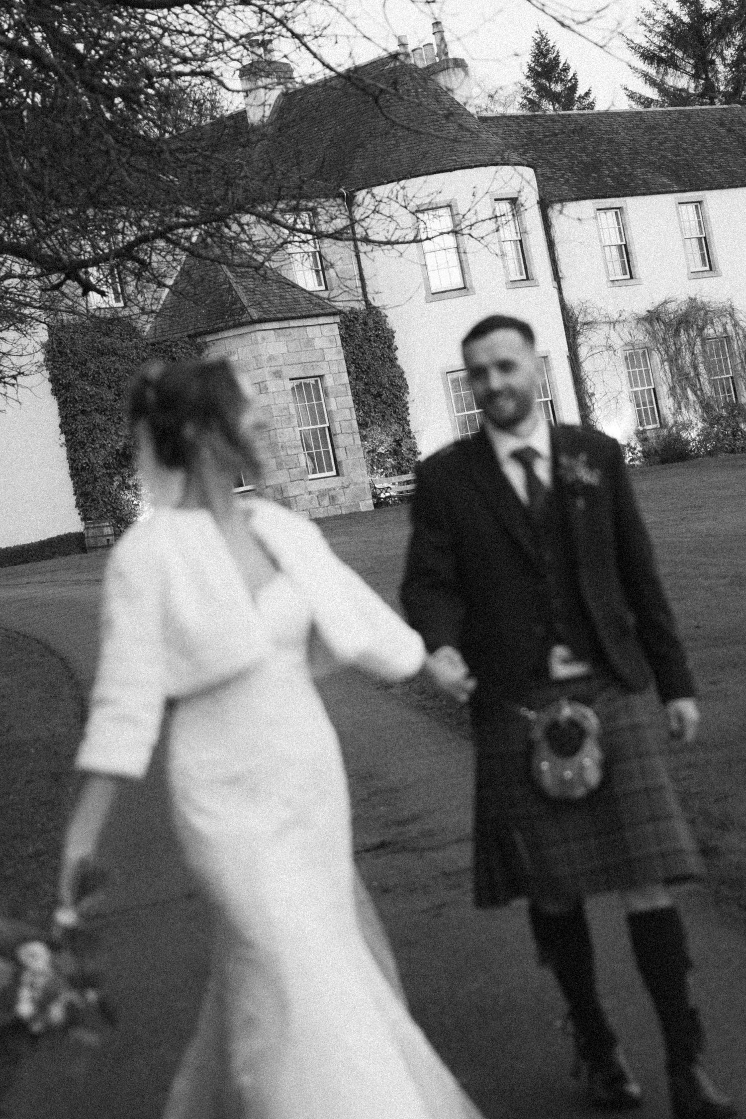 A wedding couple walks hand in hand down a path with a white mansion in the background.