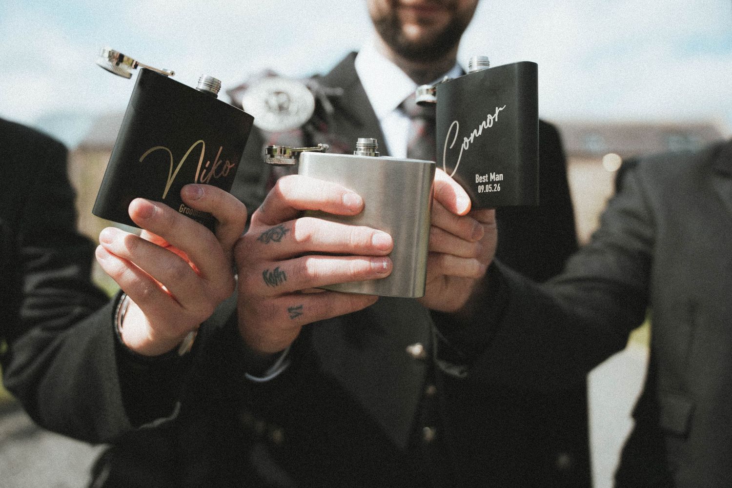 Groomsmen toasting with personalized engraved hip flasks at a wedding celebration outdoors.