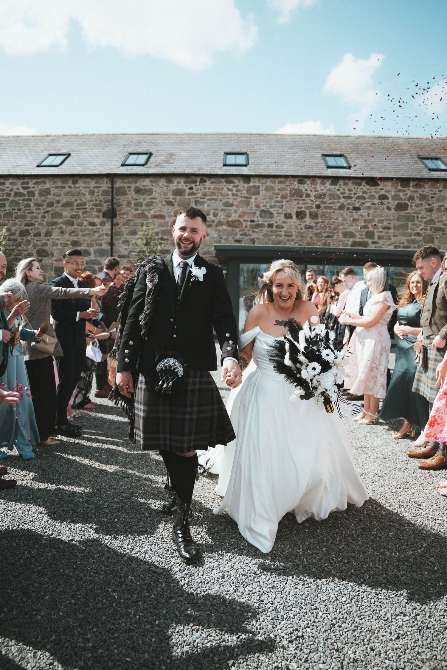 Smiling newlyweds walk through confetti shower, groom in Scottish kilt, bride in white gown, stone barn backdrop.