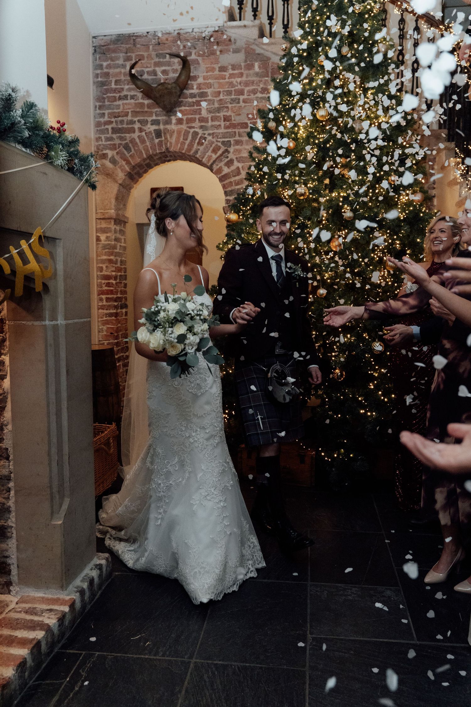 Confetti falls during winter wedding celebration with decorated Christmas tree and brick archway.