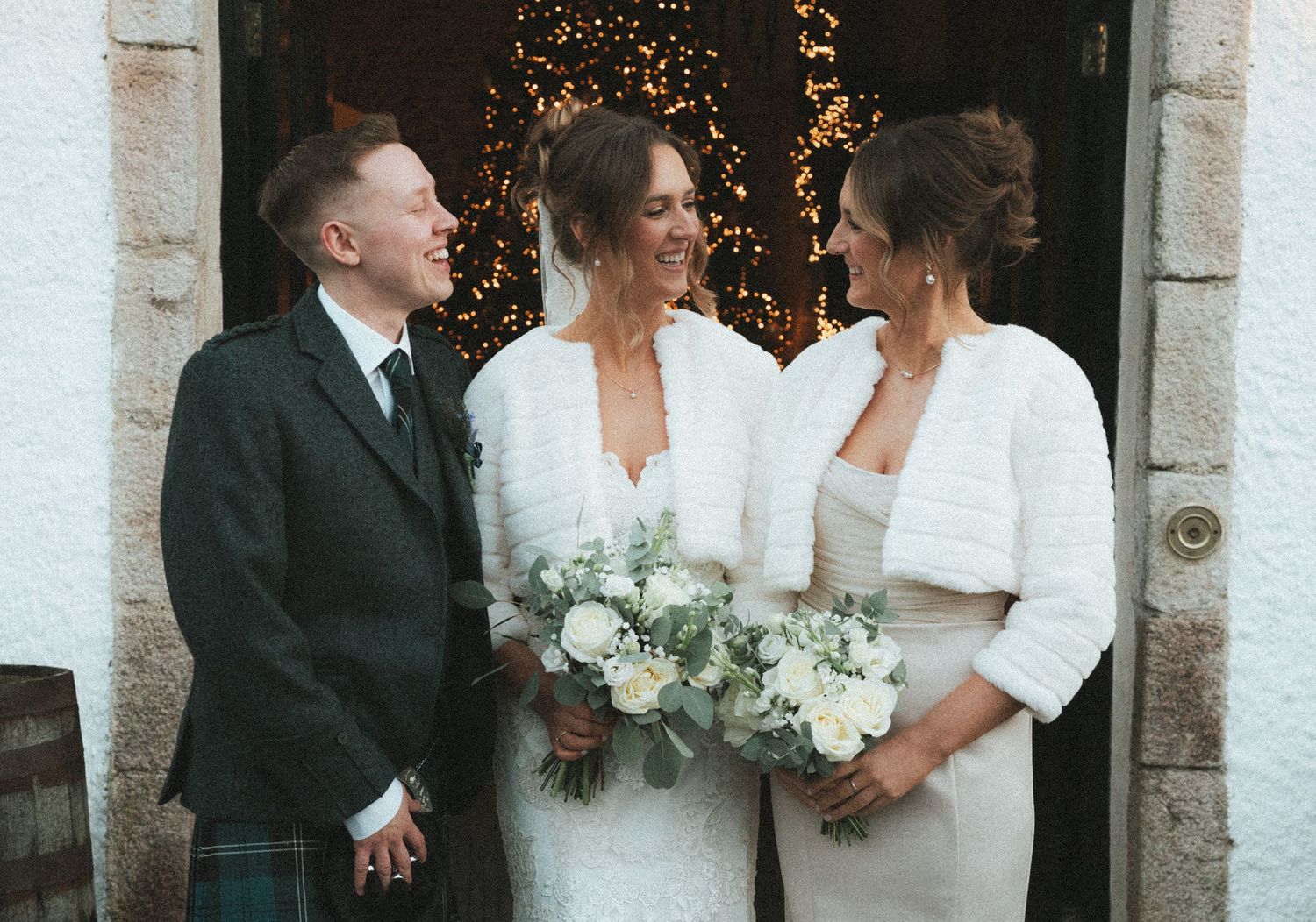 Wedding party members sharing joyful moment outside venue with festive lighting and stone architecture.