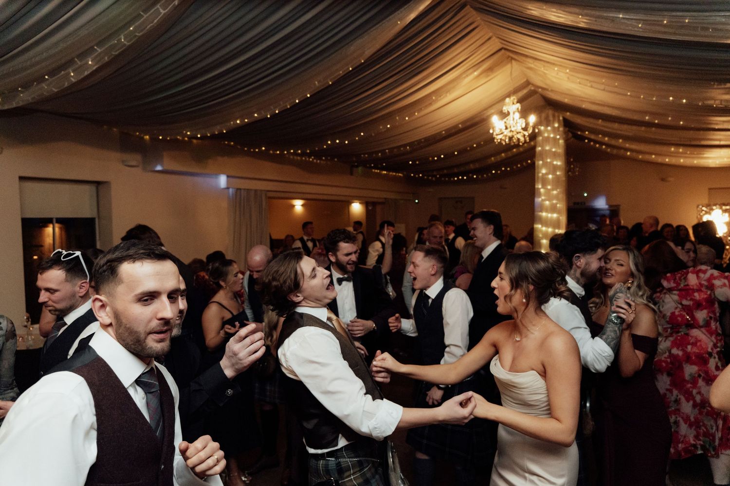 Wedding reception in full swing with guests dancing under elegant ceiling draping and chandeliers.