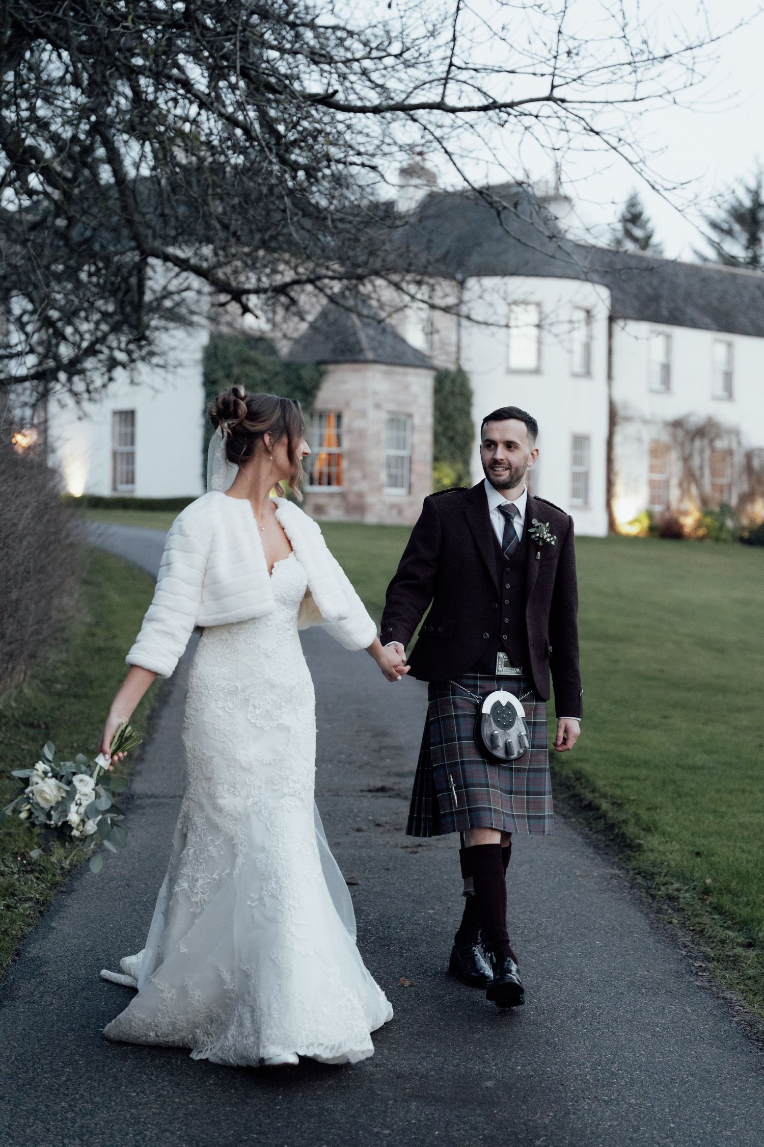 Newlyweds stroll together on a winter path near a stately white building at dusk.