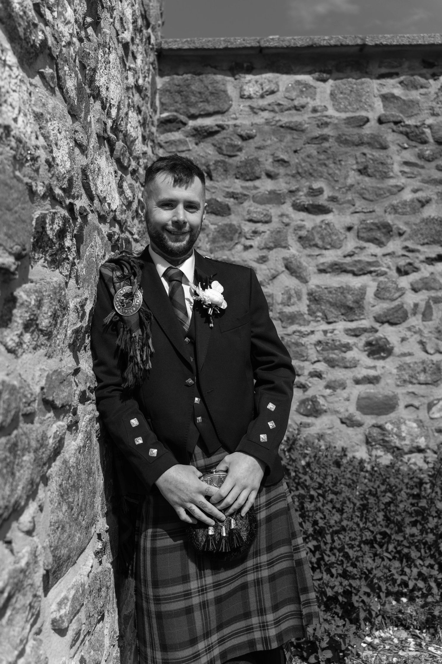 Smiling groom in traditional Scottish kilt and jacket with boutonniere leaning against stone wall in black and white photo.