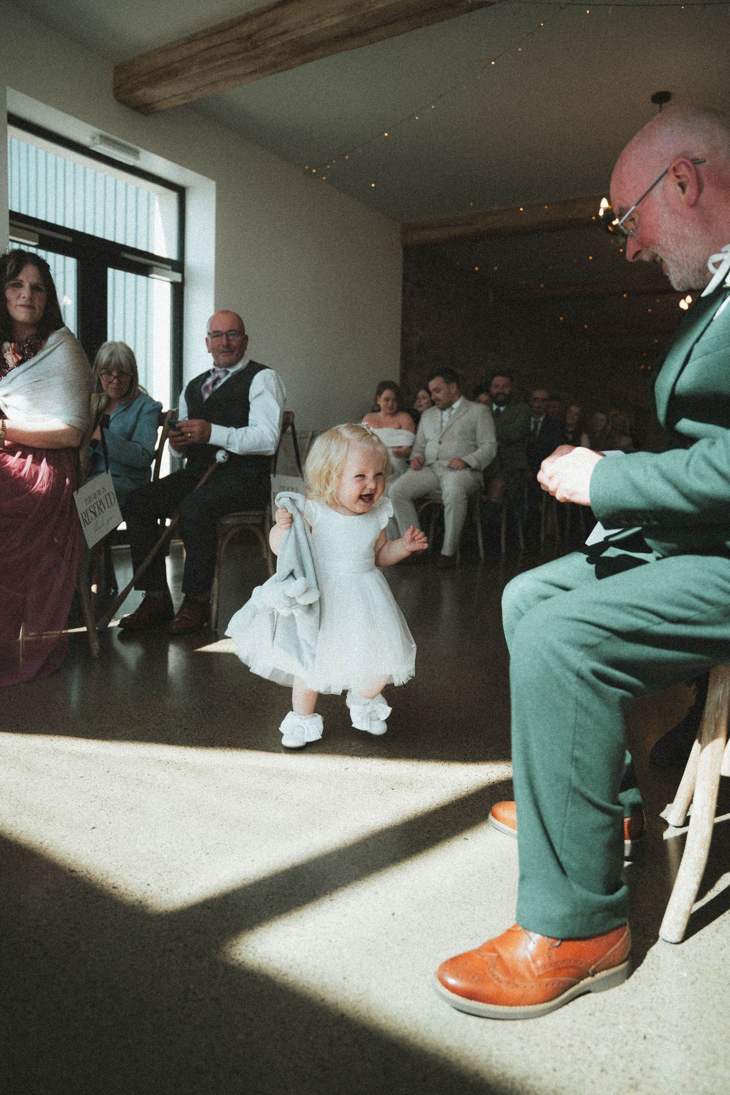 Adorable toddler flower girl in white dress walking down wedding ceremony aisle toward seated guest.