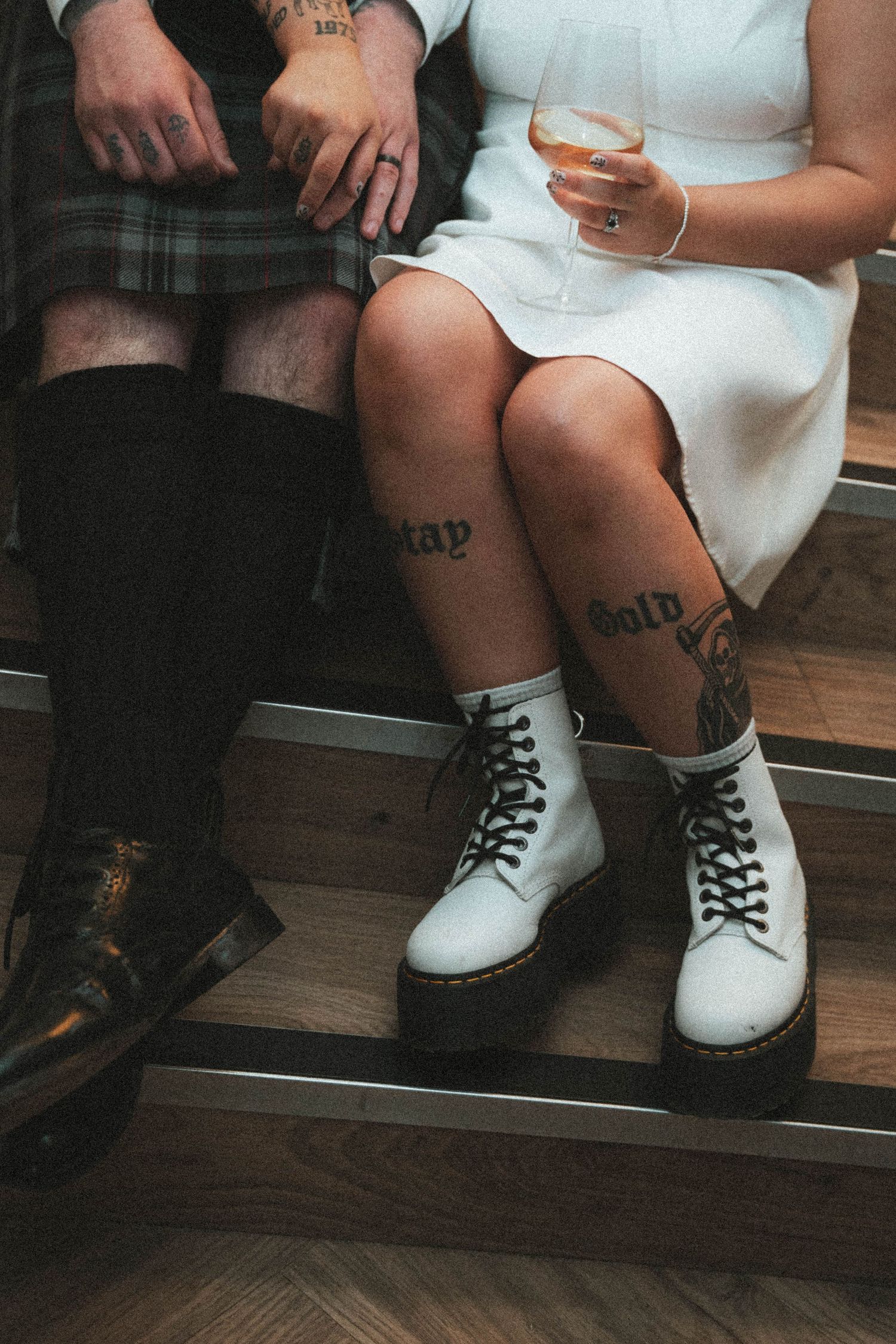 Couple with leg tattoos sit together, bride wearing white Dr Martens boots and holding rose wine glass.
