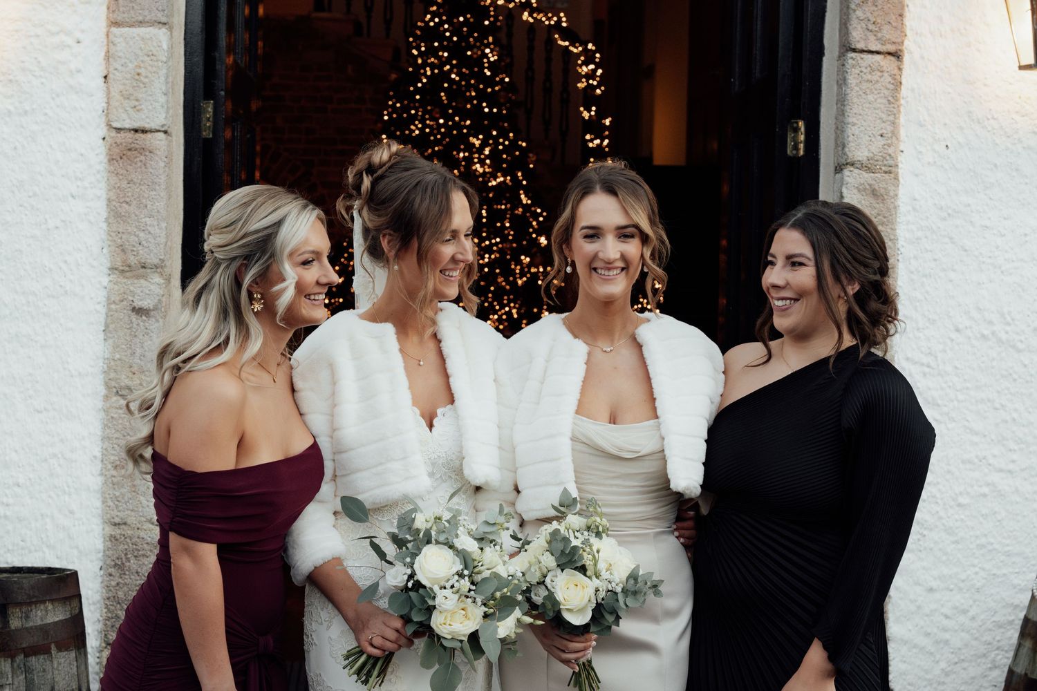 Wedding party in formal attire standing outside stone building entrance with twinkle lights and white floral bouquets.