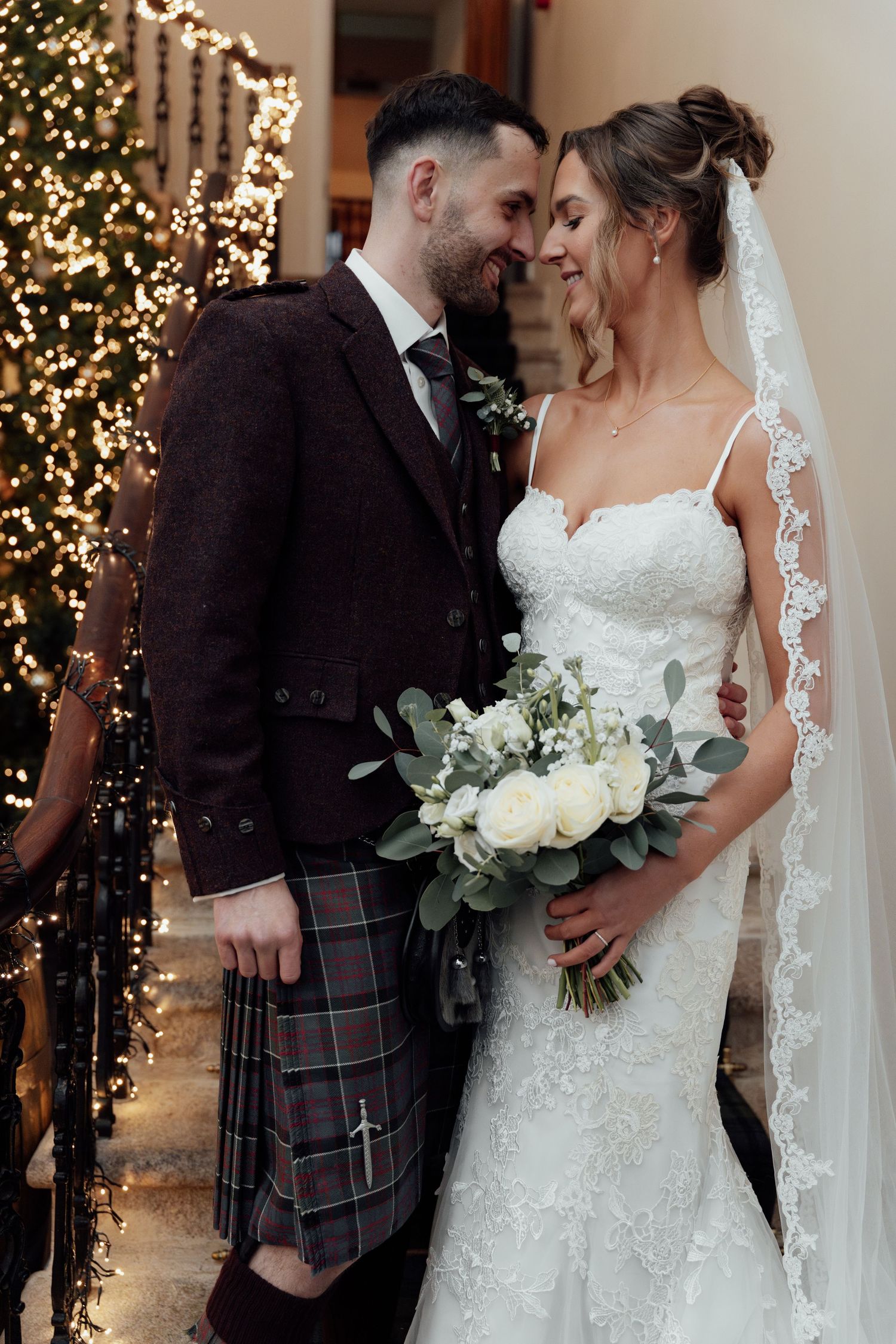 Romantic wedding portrait with Christmas lights, Scottish attire, and white floral bouquet.