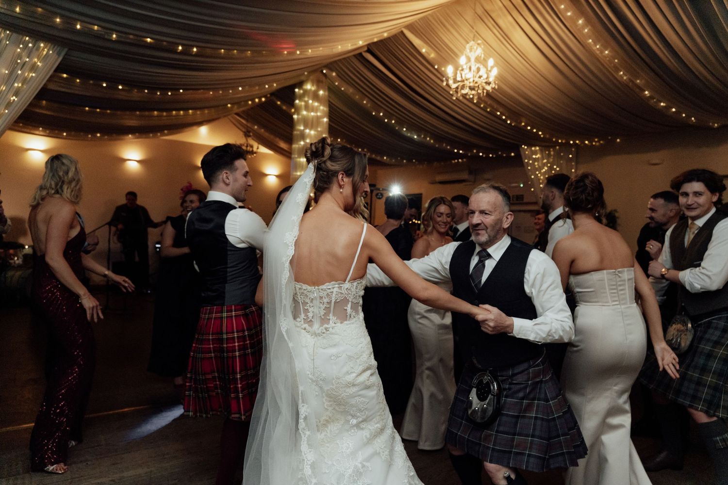 Wedding guests dance and celebrate on a dimly lit dance floor under elegant draped fabric and chandeliers.