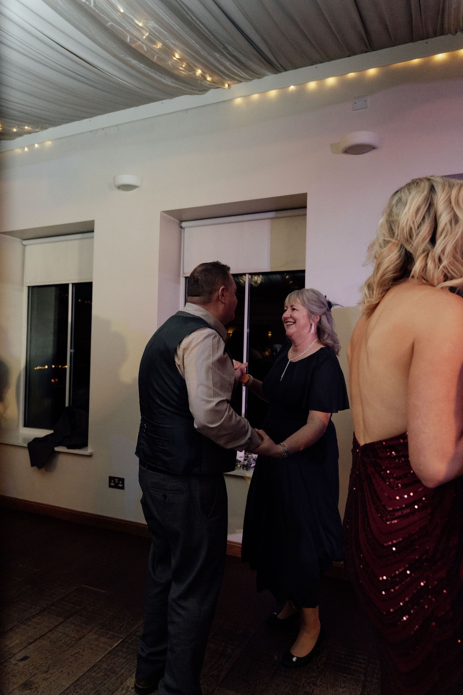 Wedding guests dancing and socializing under decorative ceiling drapes and string lights.