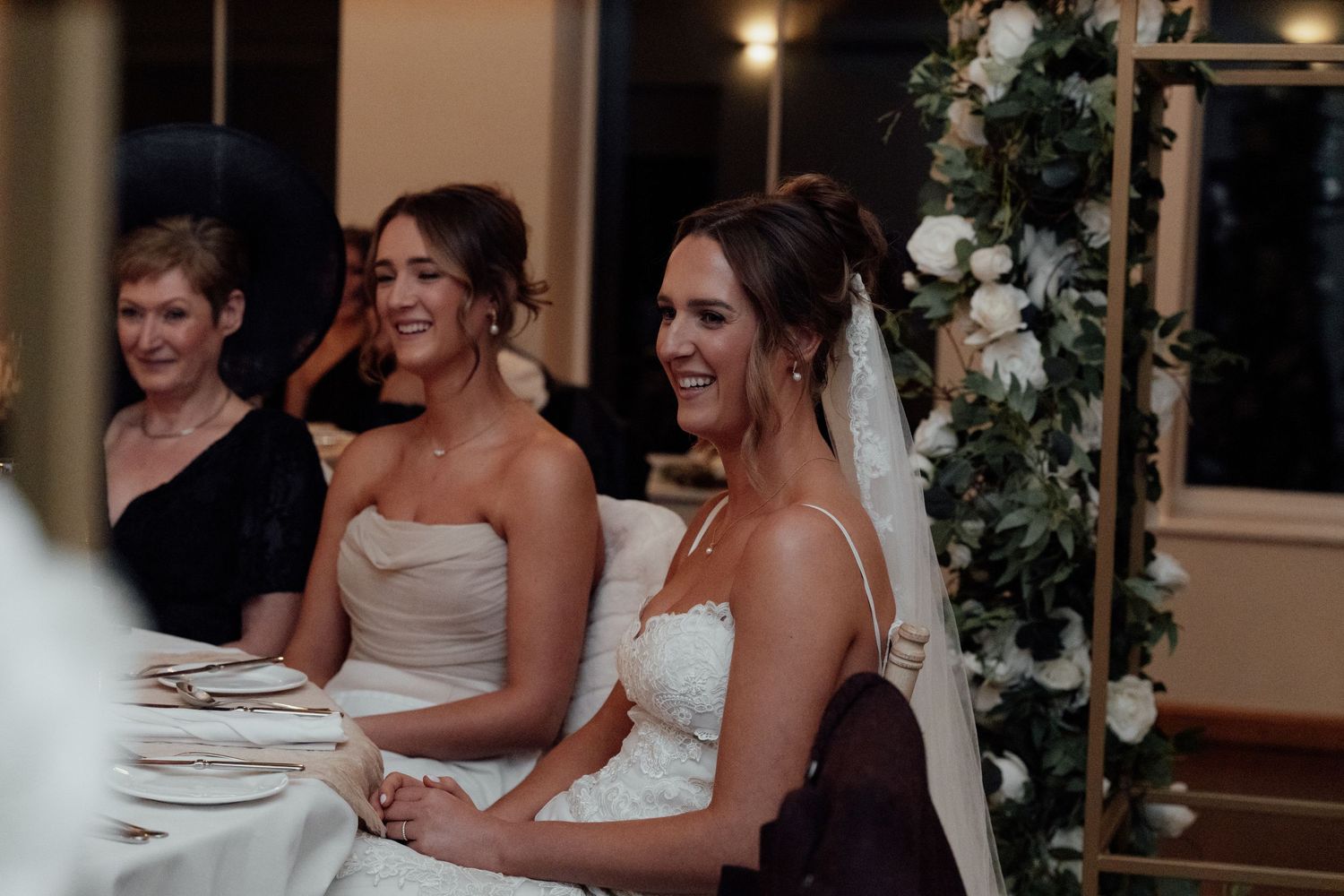 Wedding party members laugh and smile during reception dinner at decorated venue with string lights.