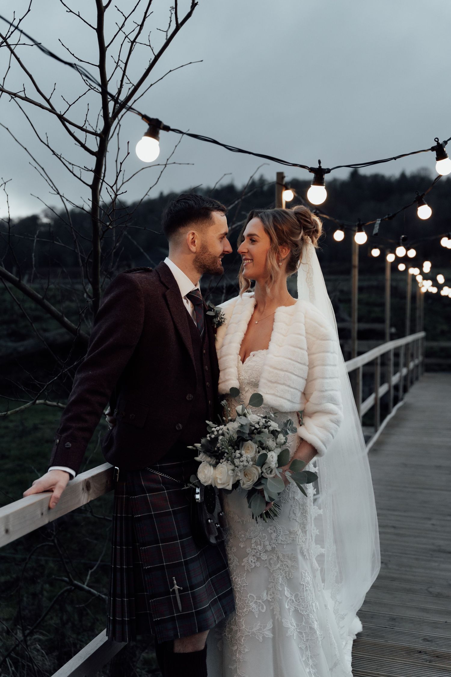 Bride and groom share romantic moment on bridge with string lights overhead.