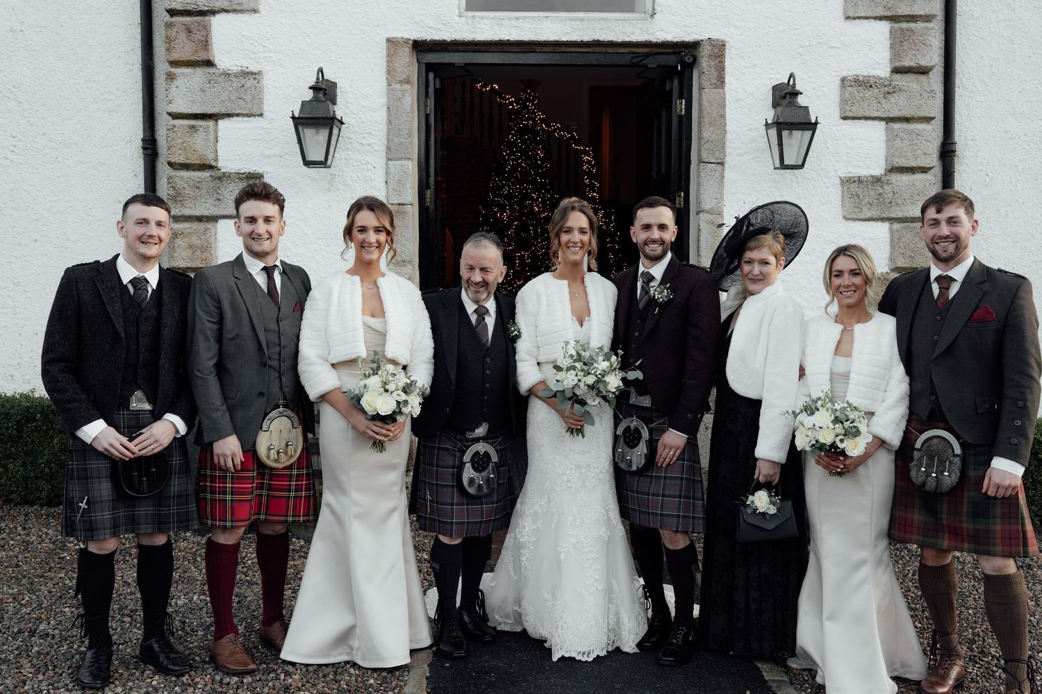 Wedding group photo in front of white building with traditional Scottish attire.
