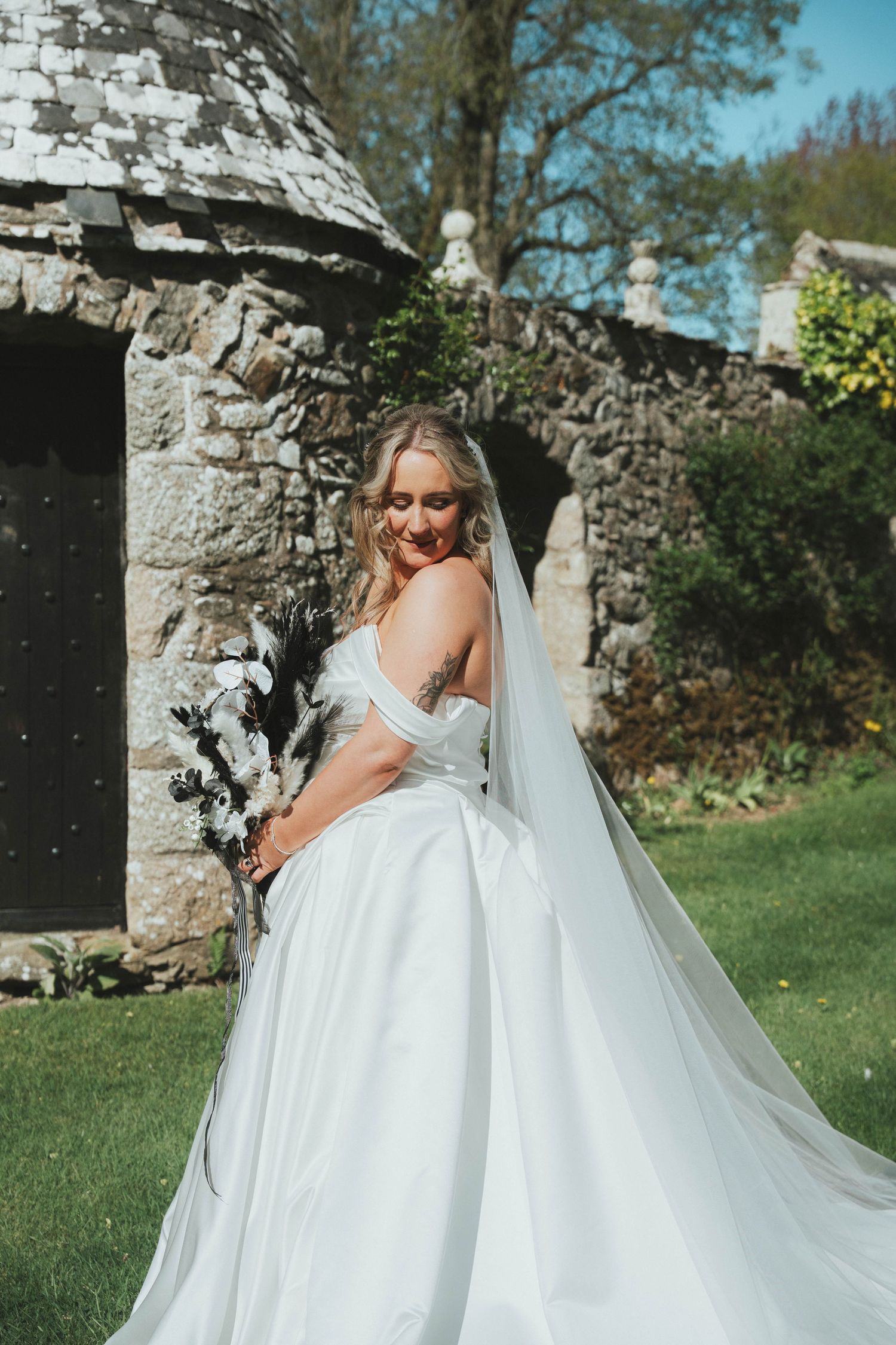 Blonde bride in flowing white off-shoulder gown with veil poses near rustic stone building in garden.