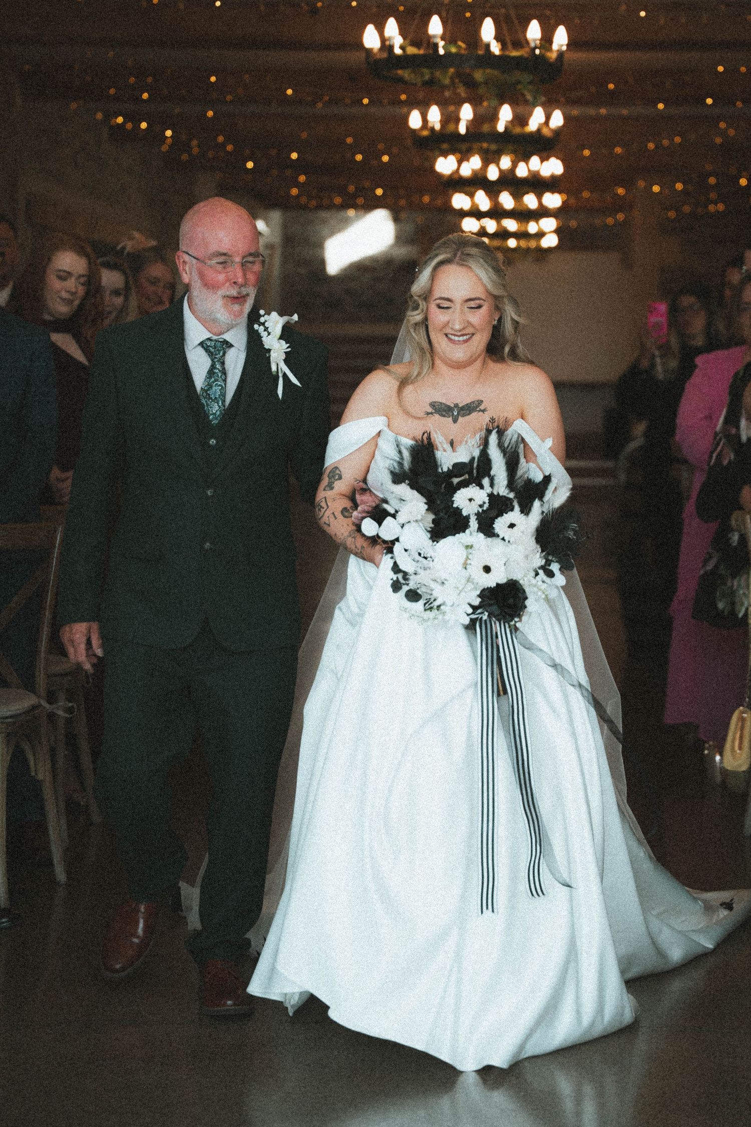 Bride in white off-shoulder gown with black bouquet walks down aisle with older gentleman at indoor wedding.