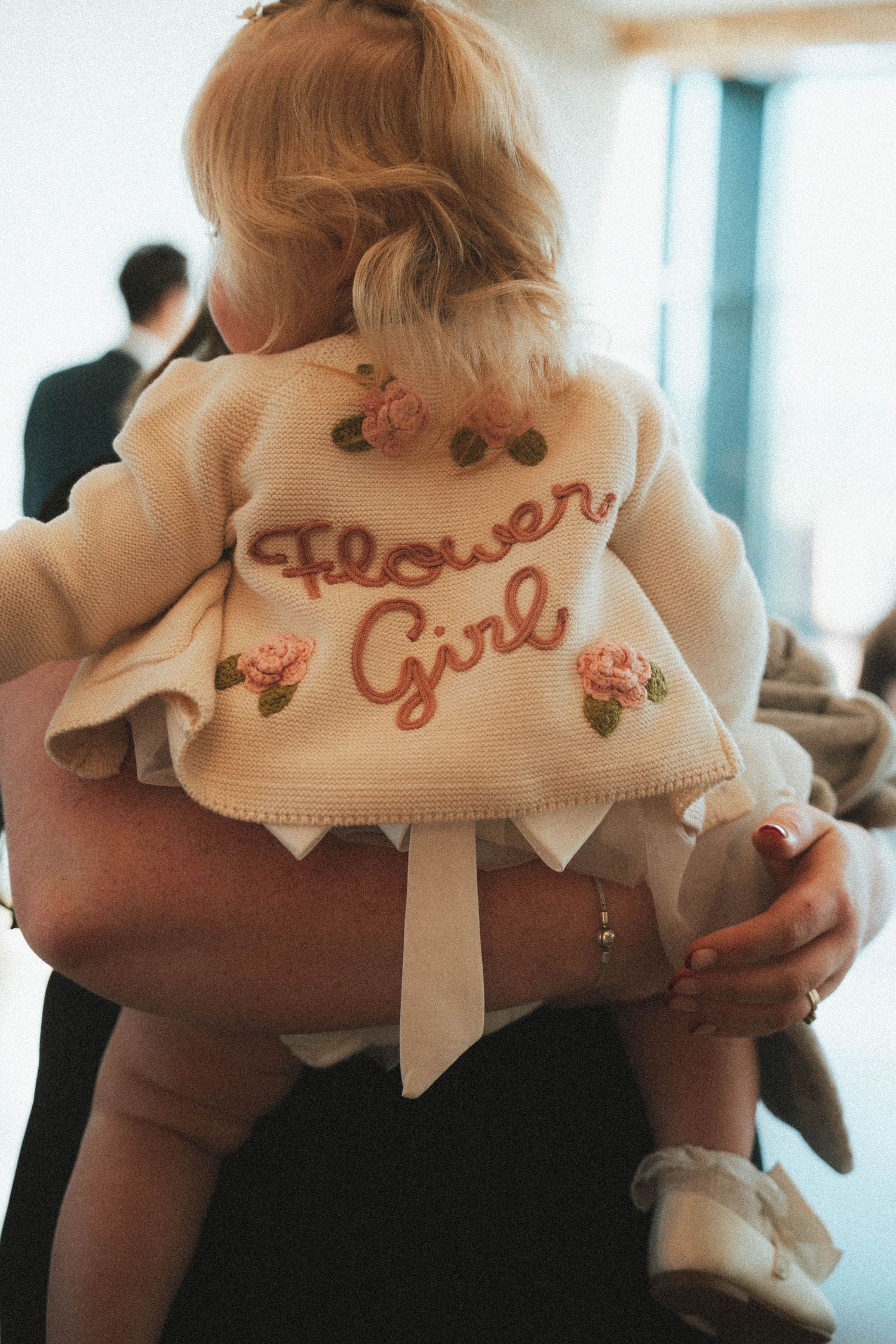 Baby flower girl in white knit cardigan embroidered with Flower Girl text being carried at wedding.