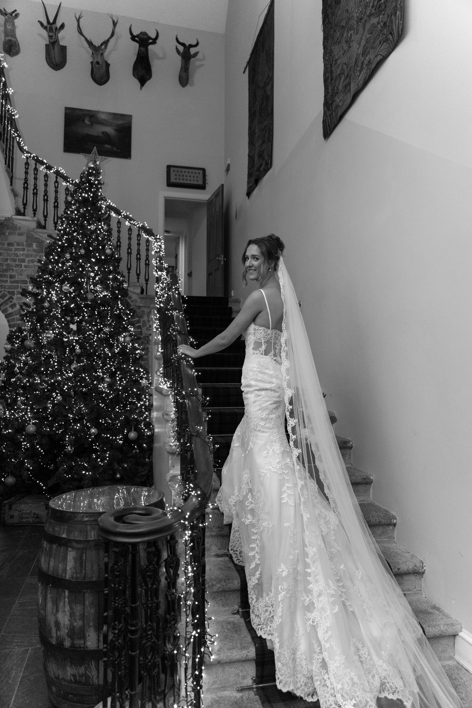 Bride in white gown poses near Christmas tree and mounted antlers in rustic indoor setting.
